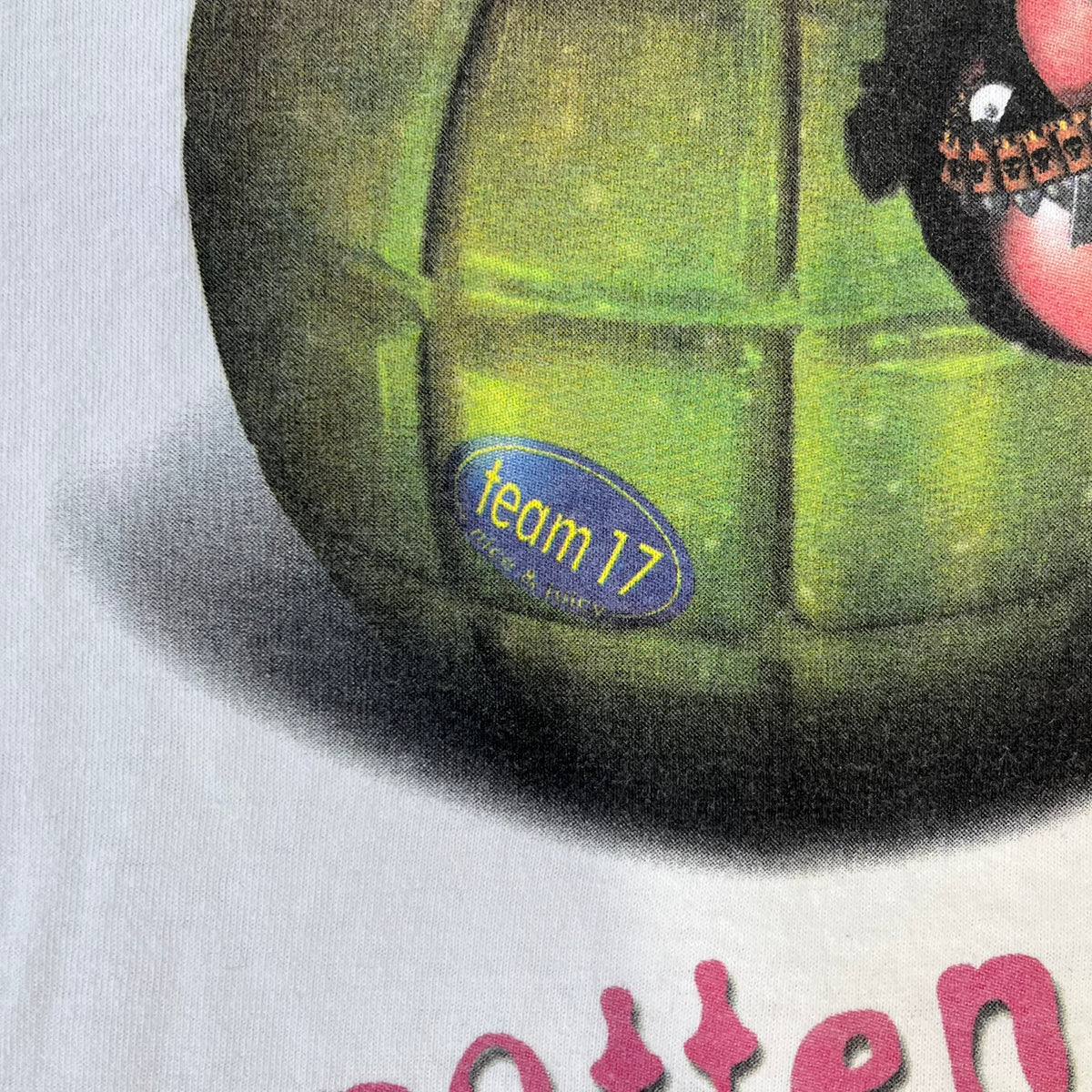 Worms 2 “Rotten 2 The Core” 1997 Tee is a white T-shirt with a front graphic of an apple styled like a grenade, with an angry worm holding a gun coming out of it. The Worms 2 logo sits to the left, and red “Rotten 2 The Core” text below the apple is followed by the website and company logos and a small 1997 licensing mark.