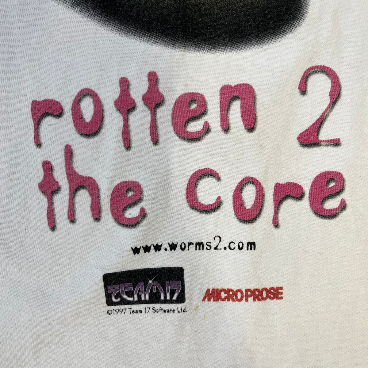 Worms 2 “Rotten 2 The Core” 1997 Tee is a white T-shirt with a front graphic of an apple styled like a grenade, with an angry worm holding a gun coming out of it. The Worms 2 logo sits to the left, and red “Rotten 2 The Core” text below the apple is followed by the website and company logos and a small 1997 licensing mark.