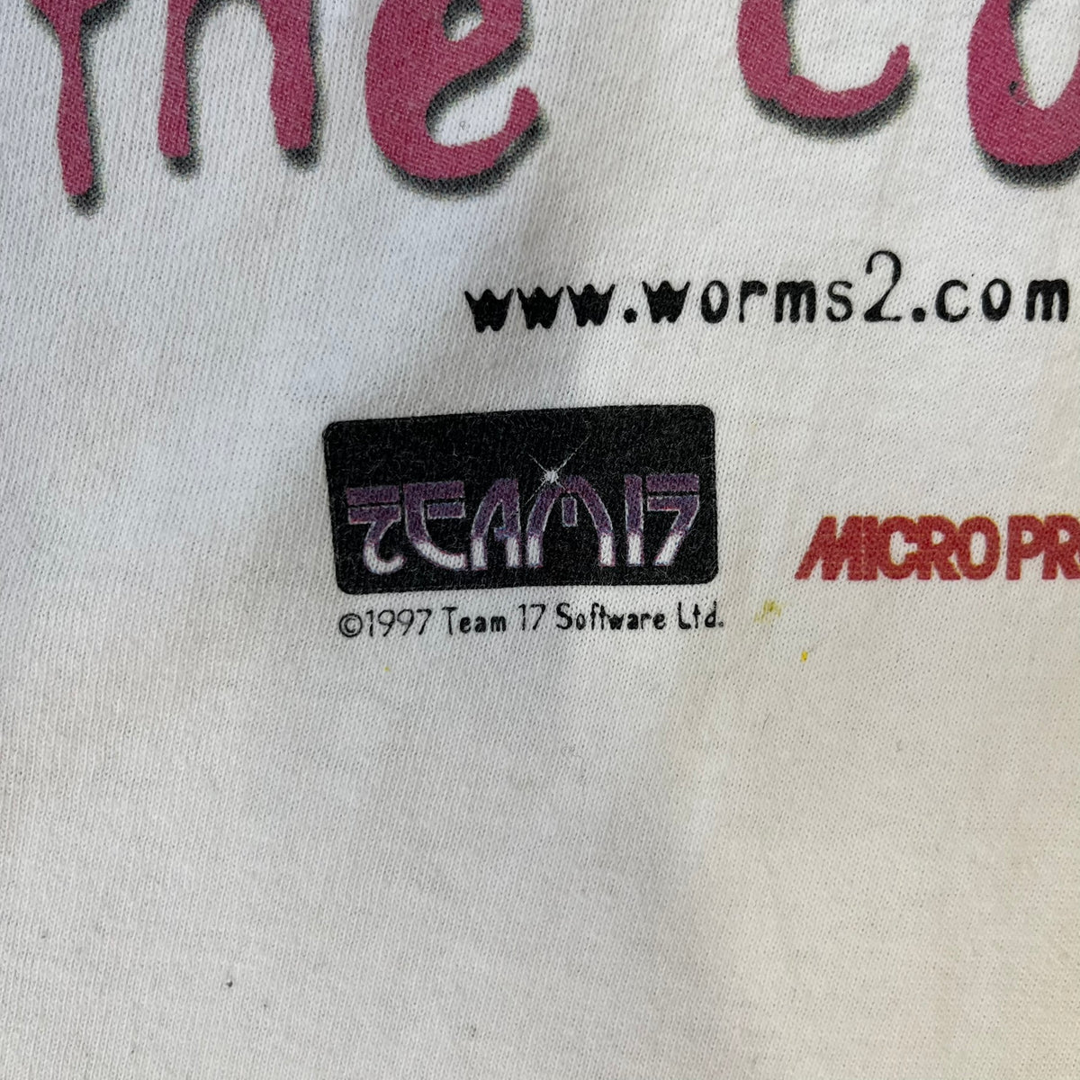 Worms 2 “Rotten 2 The Core” 1997 Tee is a white T-shirt with a front graphic of an apple styled like a grenade, with an angry worm holding a gun coming out of it. The Worms 2 logo sits to the left, and red “Rotten 2 The Core” text below the apple is followed by the website and company logos and a small 1997 licensing mark.