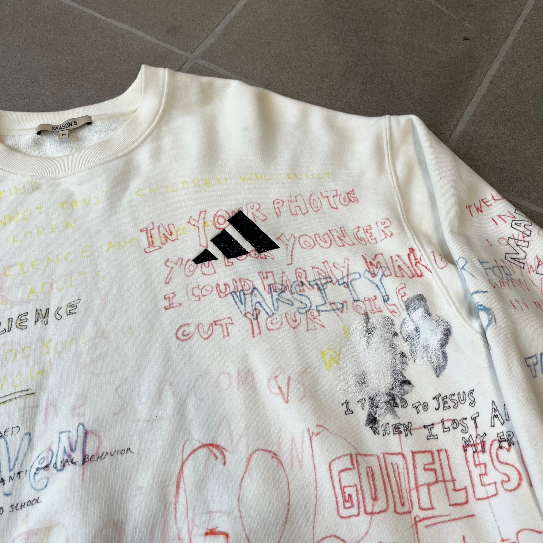 Yeezy Season 5 Scribbled Adidas Crewneck by ERD features an oversized, boxy silhouette in the Arctic colorway. The 100% cotton Adidas crewneck has handwritten-style printed graphics and phrases across the garment, including a embroidered Adidas Three Stripes logo on the chest. It was made in Italy and designed by Henri Alexander Levy from Enfants Riches Déprimés.