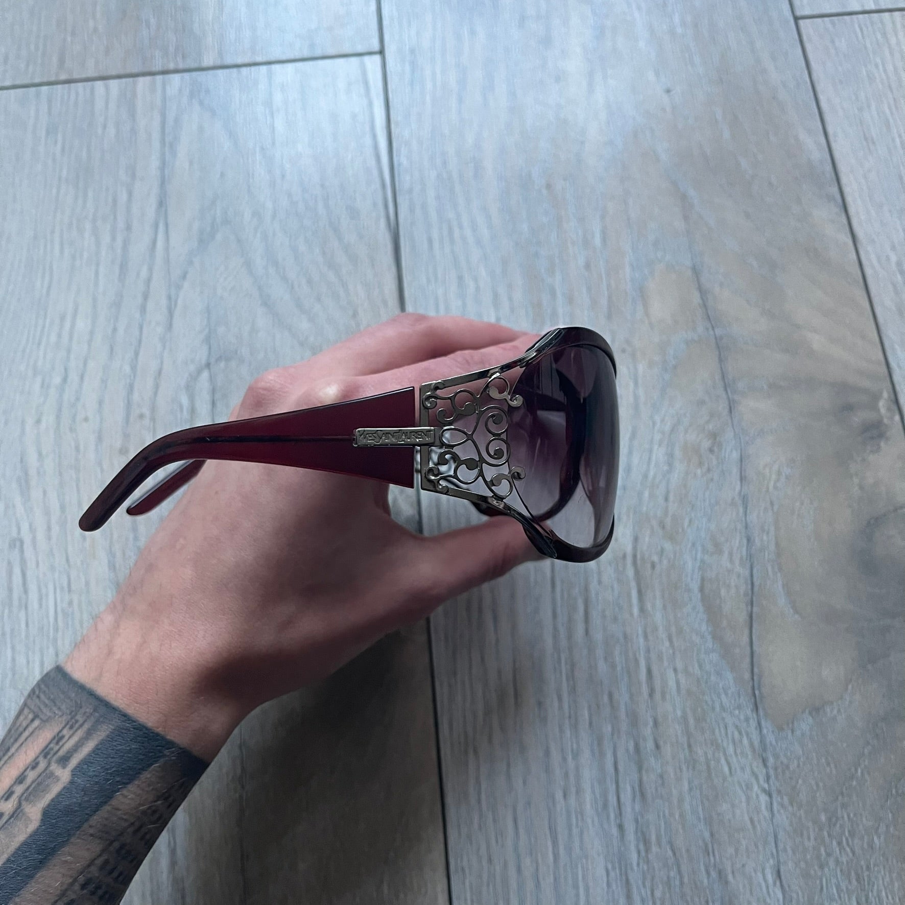 The Yves Saint Laurent 2007 Swirls 6412/S Sunglasses are an oversized model from 2007 with Category 3 sun protection. They feature oversized gradient grey lenses and burgundy (bordeaux) plastic frames, with silver-tone metal swirl details connecting the lenses to the temples. The temples are finished with a metal Yves Saint Laurent logo integrated into the swirl hardware. Made in Italy, with all original markings located on the inside. They come with a white leather YSL case.