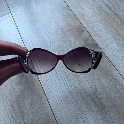 The Yves Saint Laurent 2007 Swirls 6412/S Sunglasses are an oversized model from 2007 with Category 3 sun protection. They feature oversized gradient grey lenses and burgundy (bordeaux) plastic frames, with silver-tone metal swirl details connecting the lenses to the temples. The temples are finished with a metal Yves Saint Laurent logo integrated into the swirl hardware. Made in Italy, with all original markings located on the inside. They come with a white leather YSL case.