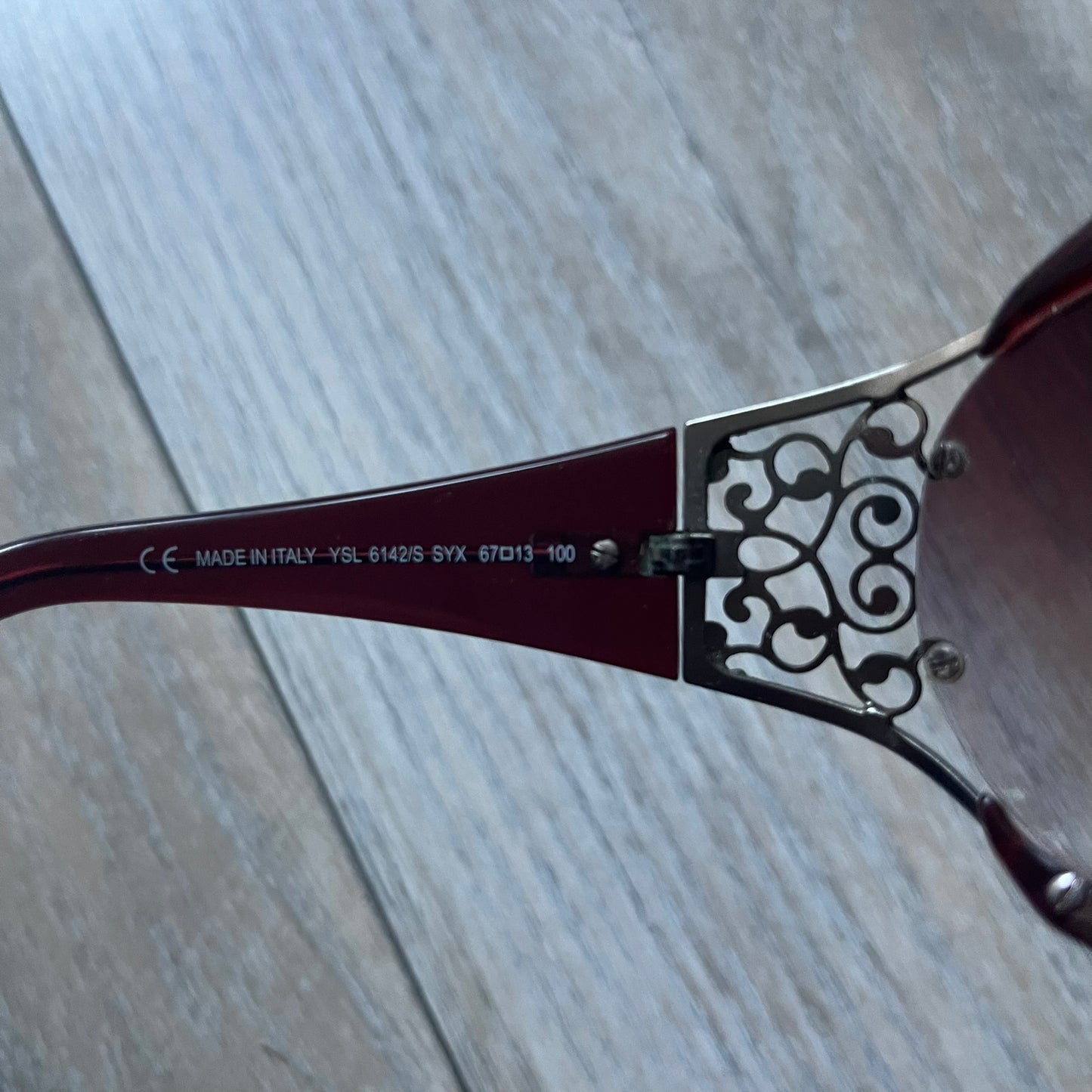 The Yves Saint Laurent 2007 Swirls 6412/S Sunglasses are an oversized model from 2007 with Category 3 sun protection. They feature oversized gradient grey lenses and burgundy (bordeaux) plastic frames, with silver-tone metal swirl details connecting the lenses to the temples. The temples are finished with a metal Yves Saint Laurent logo integrated into the swirl hardware. Made in Italy, with all original markings located on the inside. They come with a white leather YSL case.