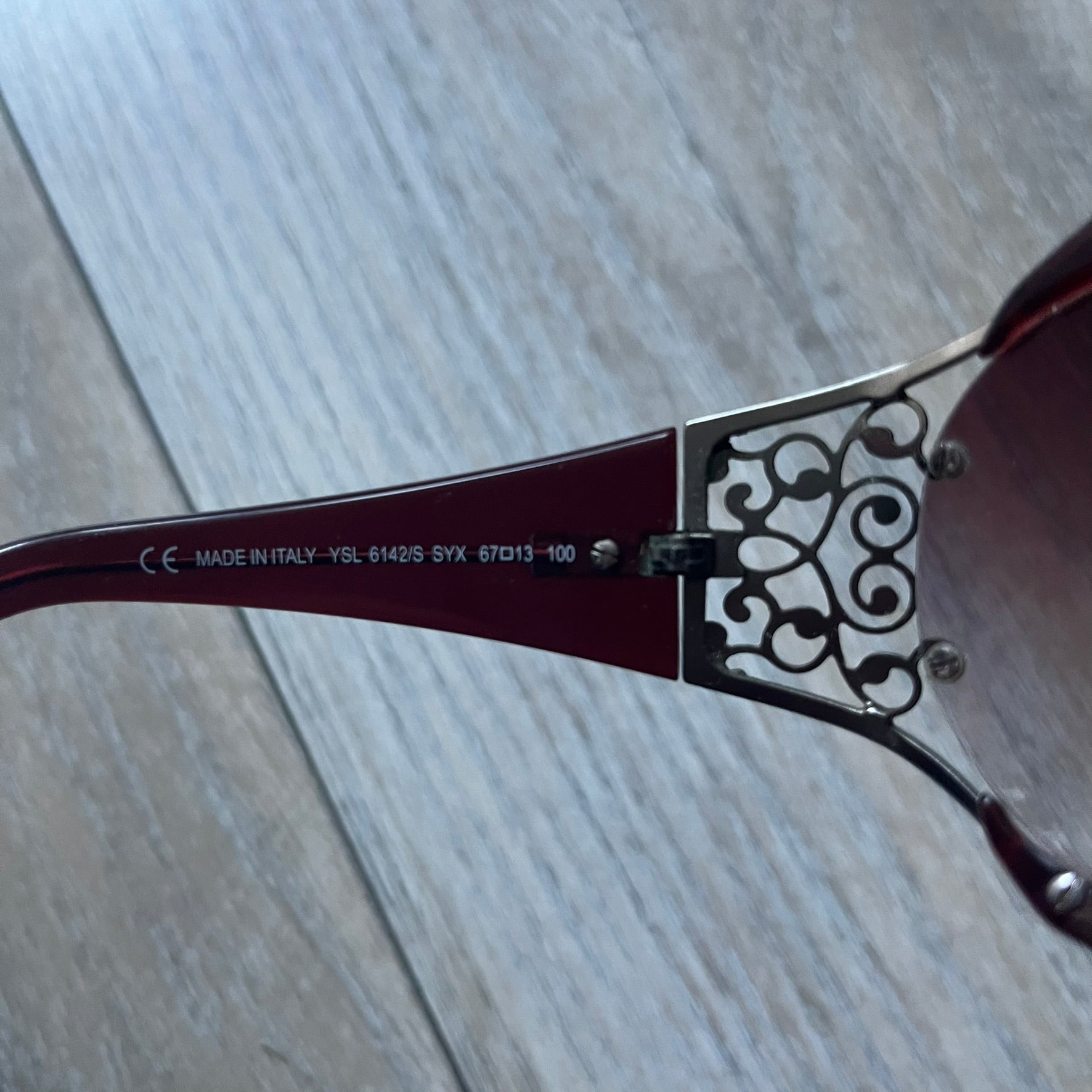 The Yves Saint Laurent 2007 Swirls 6412/S Sunglasses are an oversized model from 2007 with Category 3 sun protection. They feature oversized gradient grey lenses and burgundy (bordeaux) plastic frames, with silver-tone metal swirl details connecting the lenses to the temples. The temples are finished with a metal Yves Saint Laurent logo integrated into the swirl hardware. Made in Italy, with all original markings located on the inside. They come with a white leather YSL case.