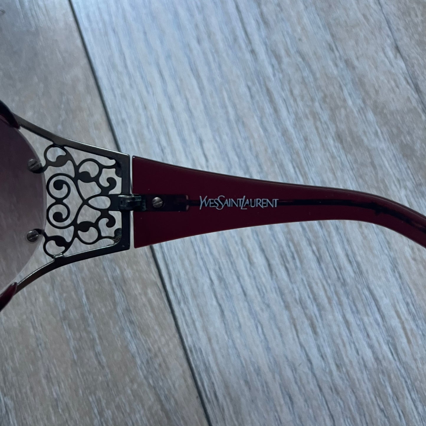 The Yves Saint Laurent 2007 Swirls 6412/S Sunglasses are an oversized model from 2007 with Category 3 sun protection. They feature oversized gradient grey lenses and burgundy (bordeaux) plastic frames, with silver-tone metal swirl details connecting the lenses to the temples. The temples are finished with a metal Yves Saint Laurent logo integrated into the swirl hardware. Made in Italy, with all original markings located on the inside. They come with a white leather YSL case.
