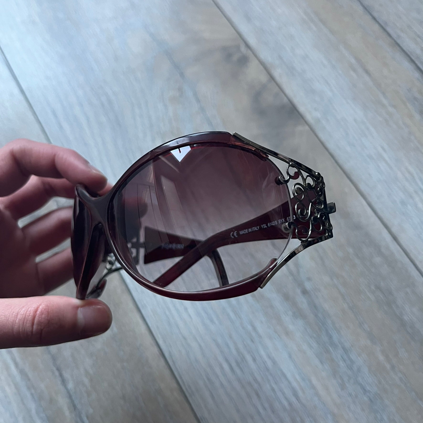 The Yves Saint Laurent 2007 Swirls 6412/S Sunglasses are an oversized model from 2007 with Category 3 sun protection. They feature oversized gradient grey lenses and burgundy (bordeaux) plastic frames, with silver-tone metal swirl details connecting the lenses to the temples. The temples are finished with a metal Yves Saint Laurent logo integrated into the swirl hardware. Made in Italy, with all original markings located on the inside. They come with a white leather YSL case.