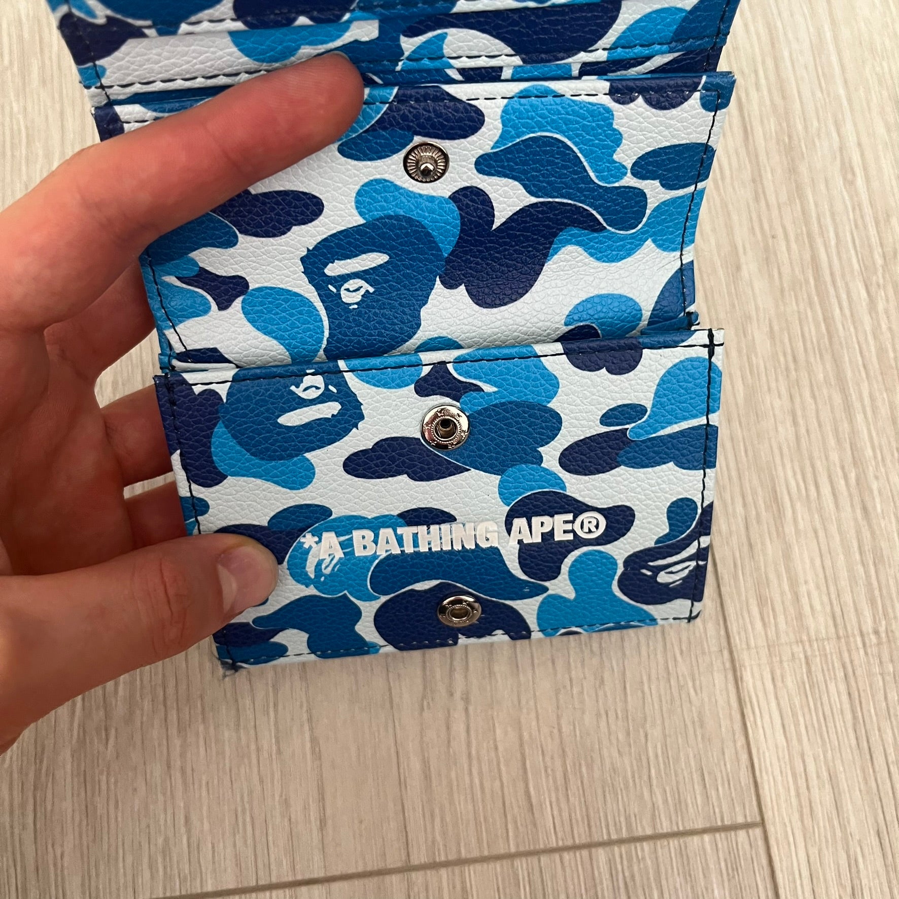 Blue and white camouflage wallet with 'A Bathing Ape' logo held by a hand on a light wooden surface.