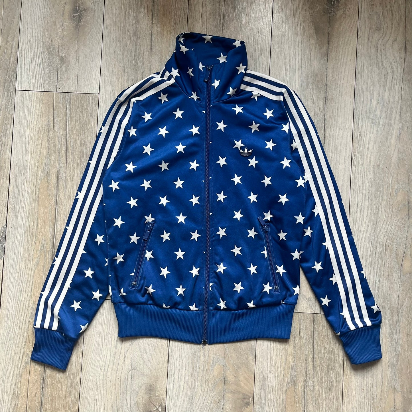 Adidas Originals Stars Blue Track Jacket is a blue zip-up track jacket featuring an all-over white star print. It includes white Adidas three-stripes along the sleeves and a small Adidas Originals patch on the chest. The design closely resembles star-pattern track jacket by Jeremy Scott for Adidas Originals.