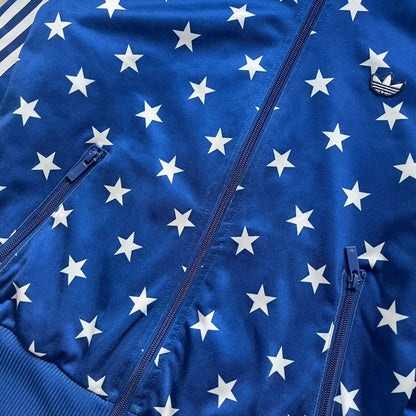 Adidas Originals Stars Blue Track Jacket is a blue zip-up track jacket featuring an all-over white star print. It includes white Adidas three-stripes along the sleeves and a small Adidas Originals patch on the chest. The design closely resembles star-pattern track jacket by Jeremy Scott for Adidas Originals.