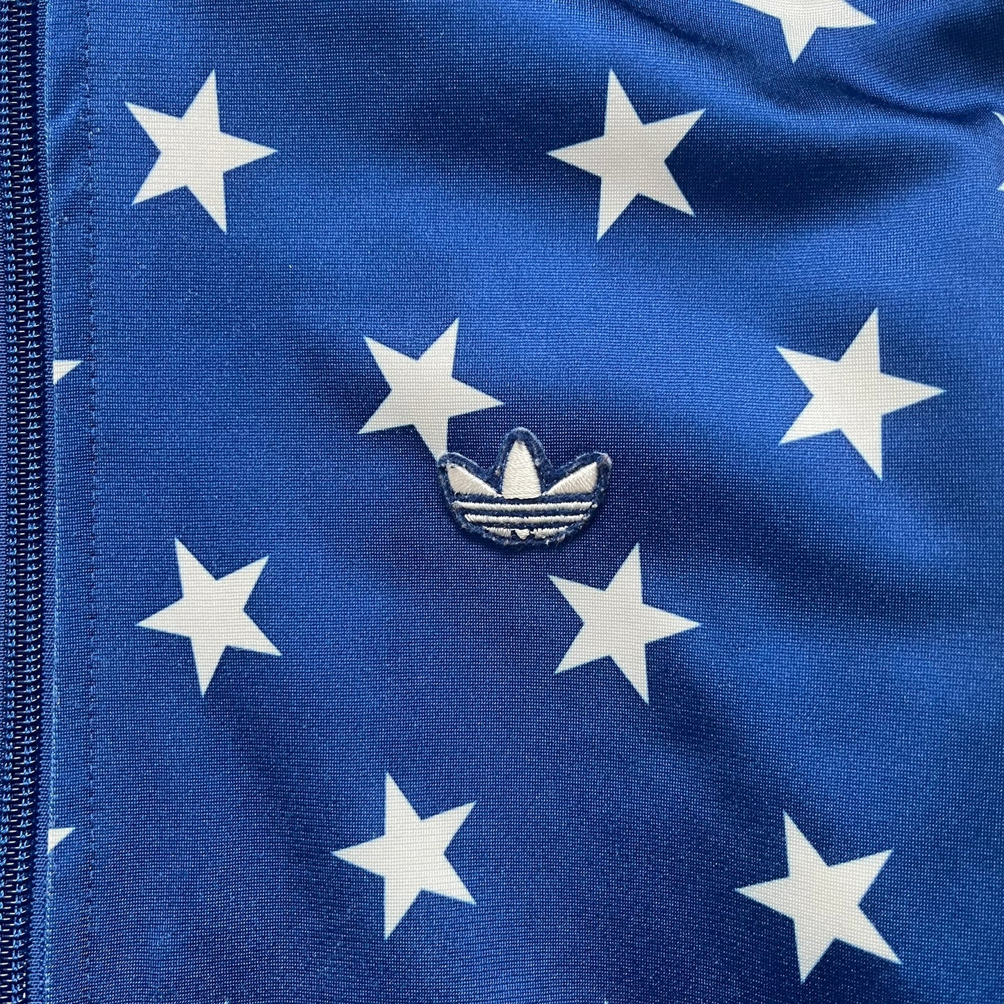 Adidas Originals Stars Blue Track Jacket is a blue zip-up track jacket featuring an all-over white star print. It includes white Adidas three-stripes along the sleeves and a small Adidas Originals patch on the chest. The design closely resembles star-pattern track jacket by Jeremy Scott for Adidas Originals.