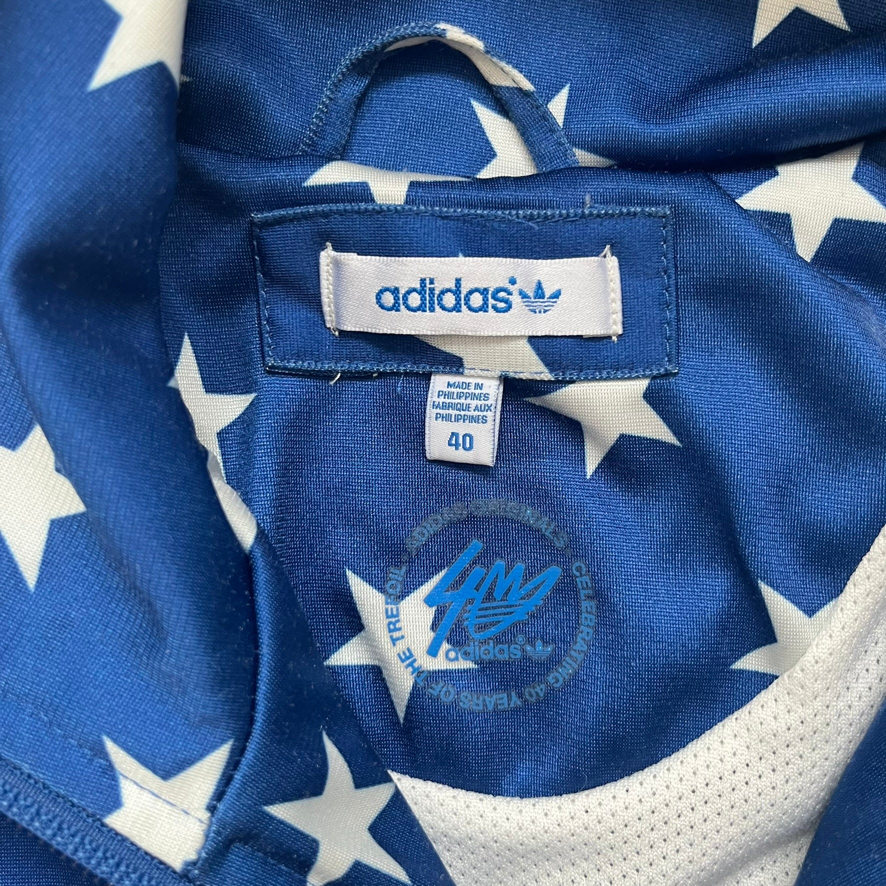 Adidas Originals Stars Blue Track Jacket is a blue zip-up track jacket featuring an all-over white star print. It includes white Adidas three-stripes along the sleeves and a small Adidas Originals patch on the chest. The design closely resembles star-pattern track jacket by Jeremy Scott for Adidas Originals.