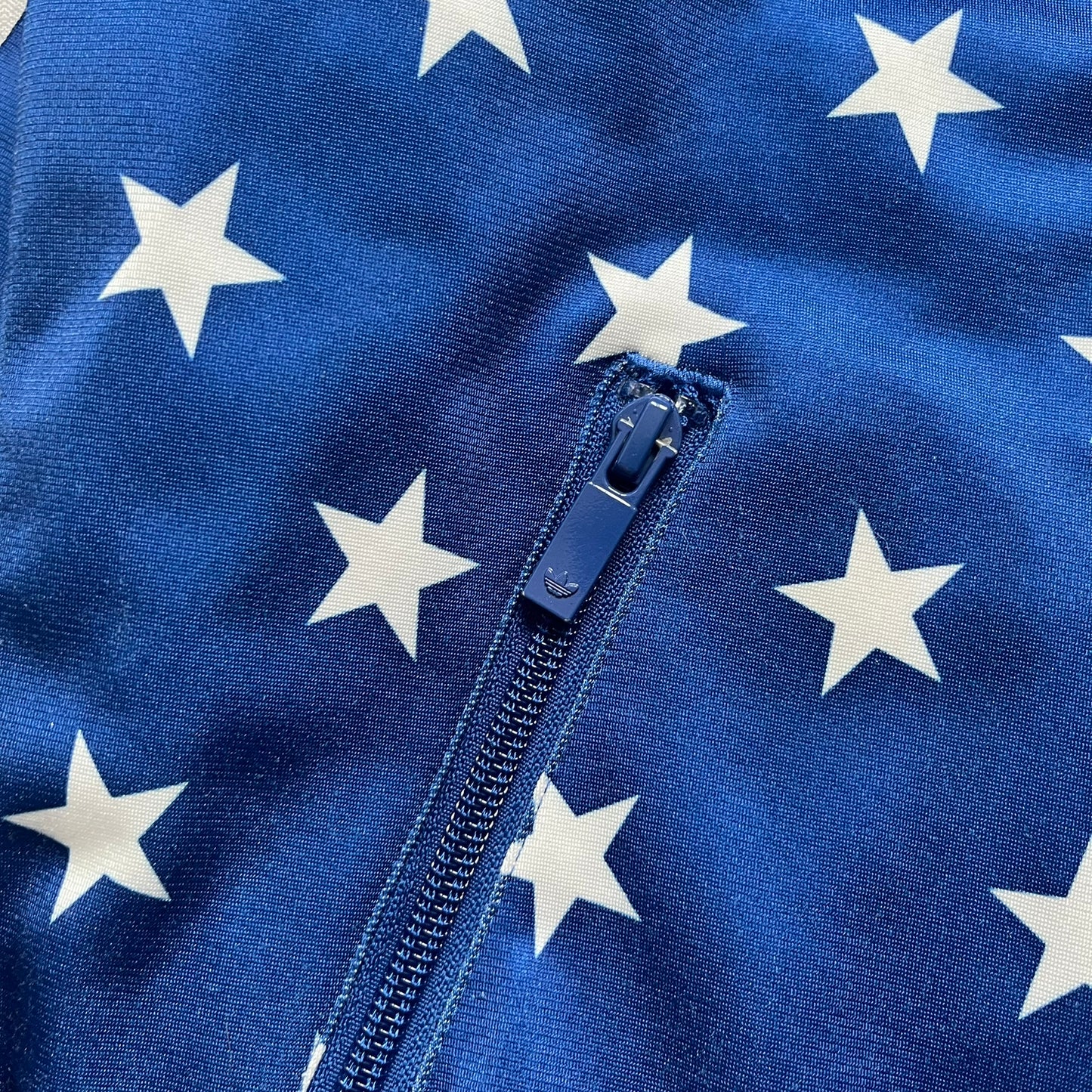 Adidas Originals Stars Blue Track Jacket is a blue zip-up track jacket featuring an all-over white star print. It includes white Adidas three-stripes along the sleeves and a small Adidas Originals patch on the chest. The design closely resembles star-pattern track jacket by Jeremy Scott for Adidas Originals.