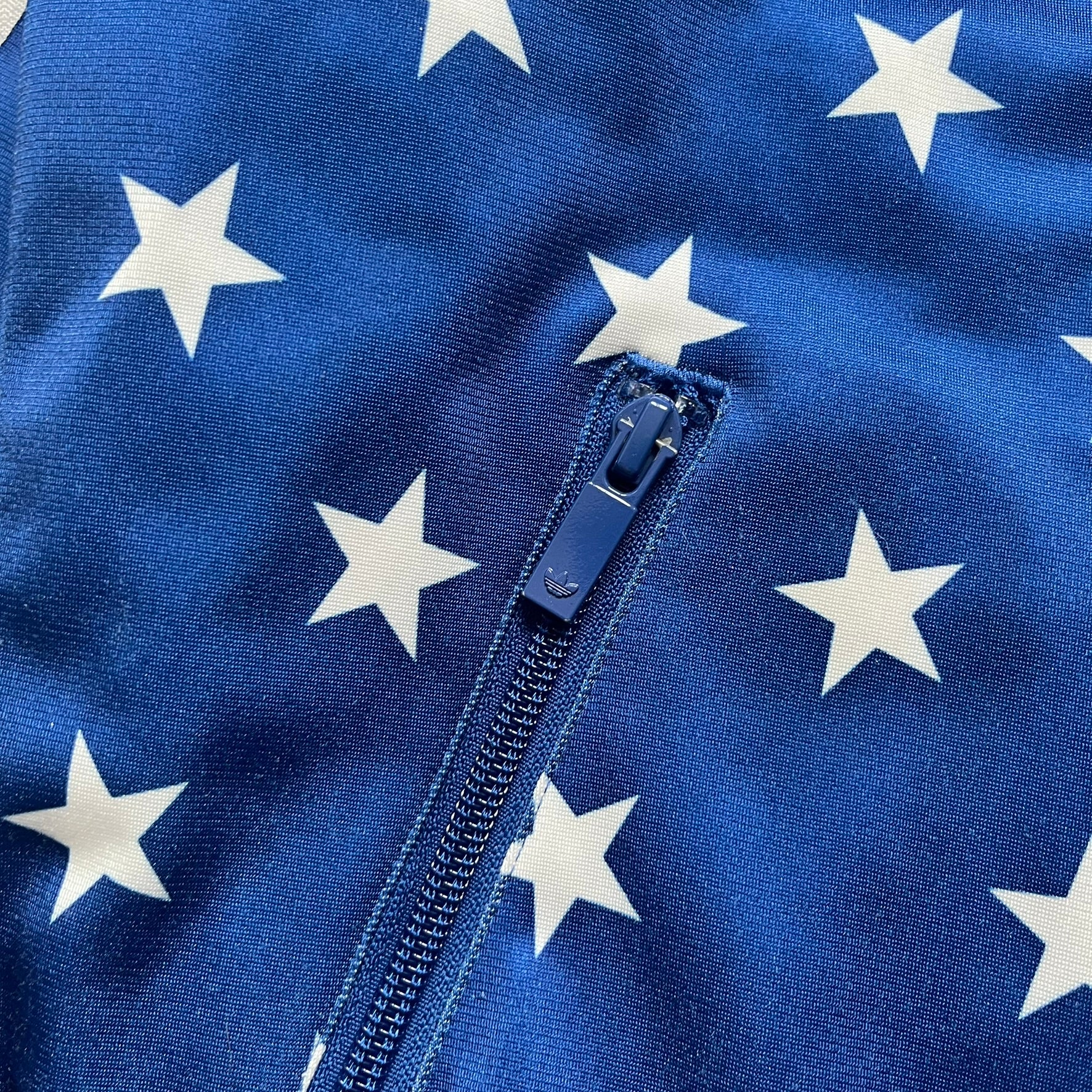 Adidas Originals Stars Blue Track Jacket is a blue zip-up track jacket featuring an all-over white star print. It includes white Adidas three-stripes along the sleeves and a small Adidas Originals patch on the chest. The design closely resembles star-pattern track jacket by Jeremy Scott for Adidas Originals.