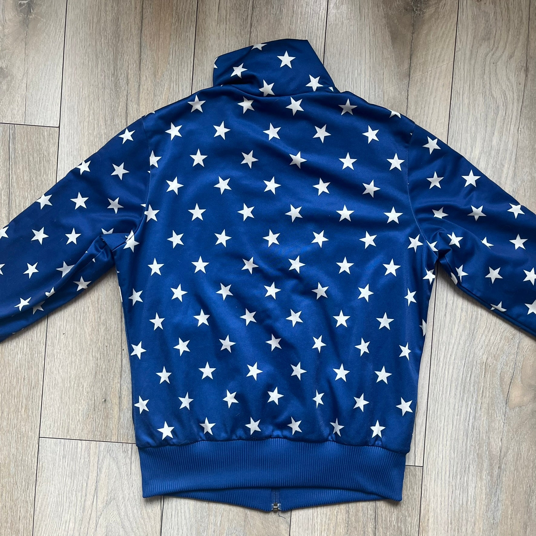 Adidas Originals Stars Blue Track Jacket is a blue zip-up track jacket featuring an all-over white star print. It includes white Adidas three-stripes along the sleeves and a small Adidas Originals patch on the chest. The design closely resembles star-pattern track jacket by Jeremy Scott for Adidas Originals.
