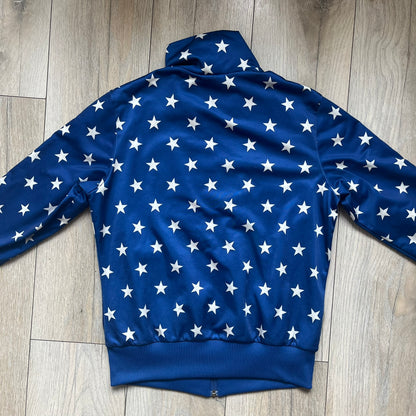 Adidas Originals Stars Blue Track Jacket is a blue zip-up track jacket featuring an all-over white star print. It includes white Adidas three-stripes along the sleeves and a small Adidas Originals patch on the chest. The design closely resembles star-pattern track jacket by Jeremy Scott for Adidas Originals.