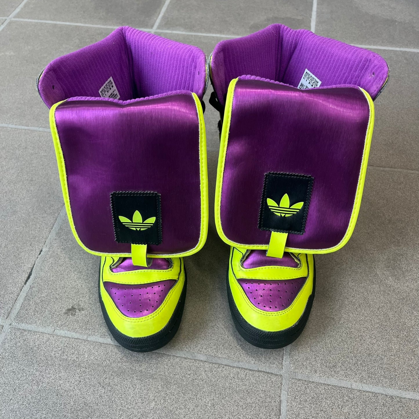 adidas Originals by Jeremy Scott 2014 JS Tall Boy Summer Sneakers feature a yellow and metallic purple high-top construction with a patent-effect synthetic upper. The design has an oversized foldable tongue, ribbed lining, ankle zipper, and abrasion-resistant rubber sole. Part of the adidas Originals by Jeremy Scott SS14 collection.
