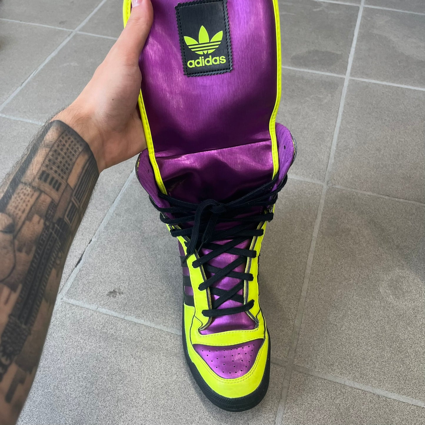 adidas Originals by Jeremy Scott 2014 JS Tall Boy Summer Sneakers feature a yellow and metallic purple high-top construction with a patent-effect synthetic upper. The design has an oversized foldable tongue, ribbed lining, ankle zipper, and abrasion-resistant rubber sole. Part of the adidas Originals by Jeremy Scott SS14 collection.
