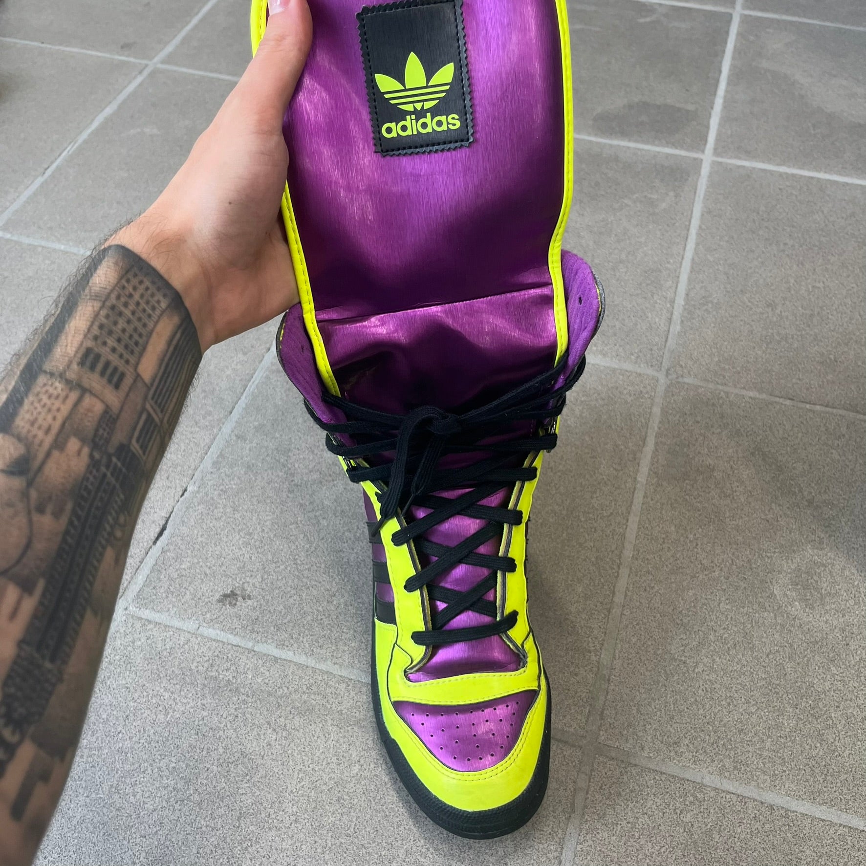 adidas Originals by Jeremy Scott 2014 JS Tall Boy Summer Sneakers feature a yellow and metallic purple high-top construction with a patent-effect synthetic upper. The design has an oversized foldable tongue, ribbed lining, ankle zipper, and abrasion-resistant rubber sole. Part of the adidas Originals by Jeremy Scott SS14 collection.
