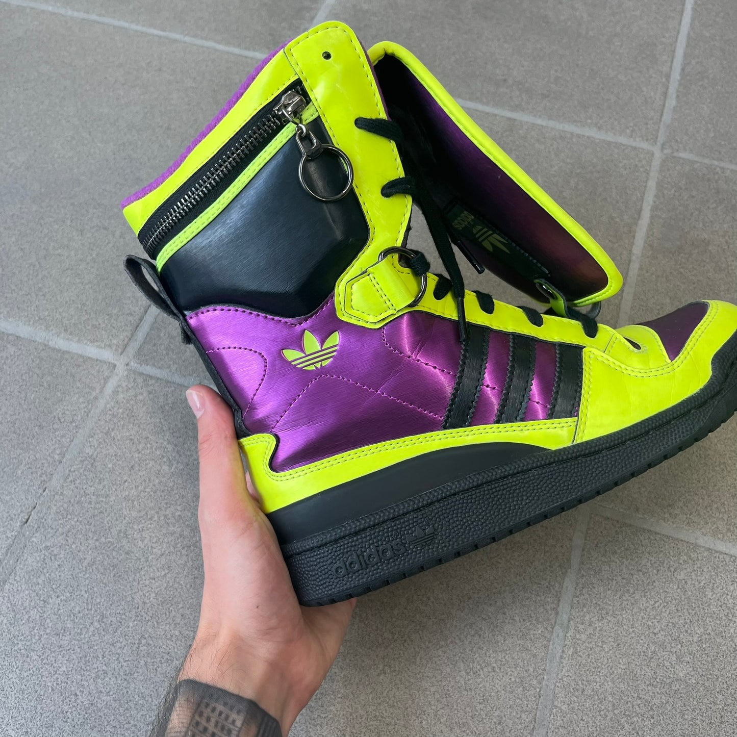 adidas Originals by Jeremy Scott 2014 JS Tall Boy Summer Sneakers feature a yellow and metallic purple high-top construction with a patent-effect synthetic upper. The design has an oversized foldable tongue, ribbed lining, ankle zipper, and abrasion-resistant rubber sole. Part of the adidas Originals by Jeremy Scott SS14 collection.
