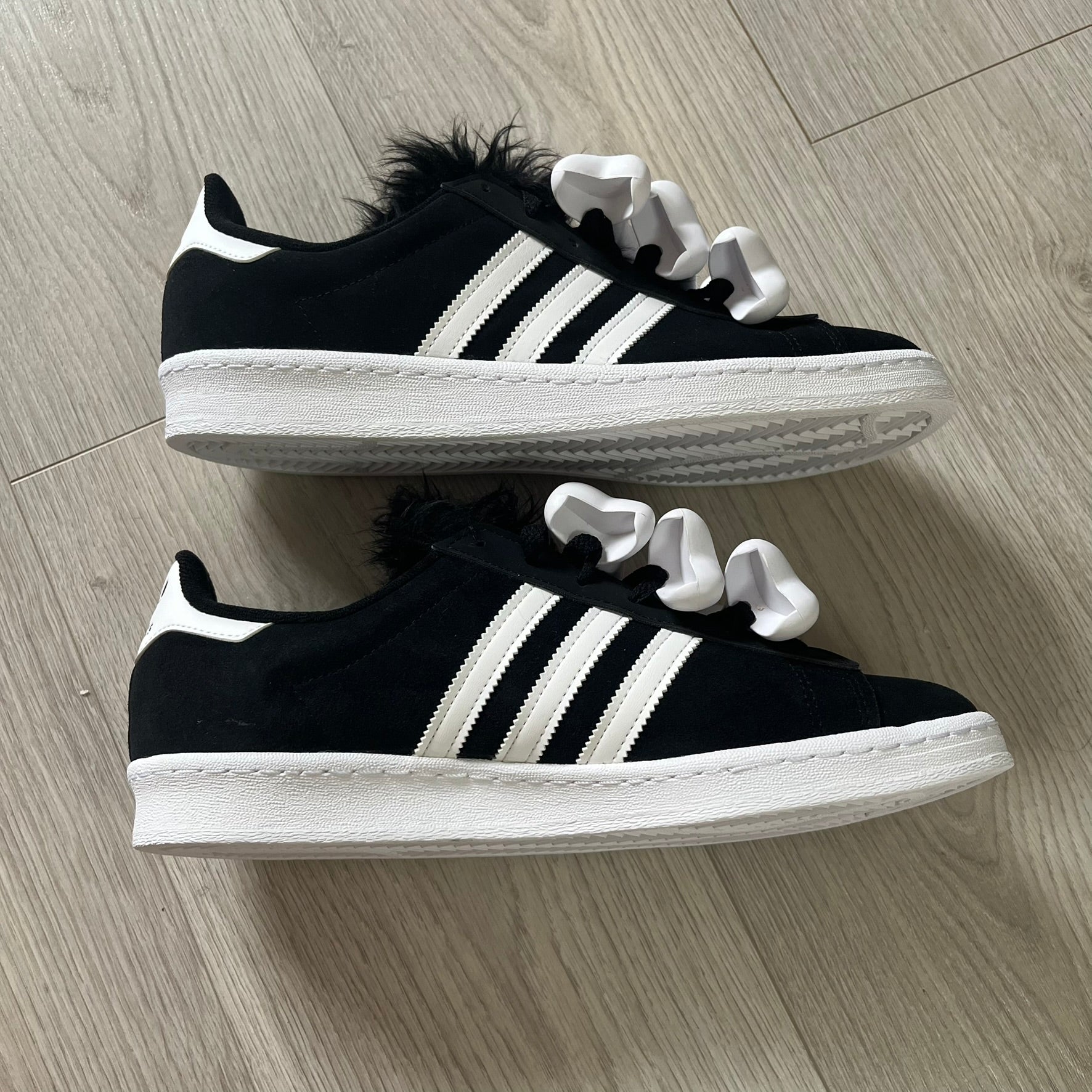 jeremy scott’s take on the adidas campus 80s includes black leather, serrated stripes, and unique faux fur tongues. detachable white bones on the laces add a customizable, standout touch. this 2023 release balances classic style with avant-garde details.