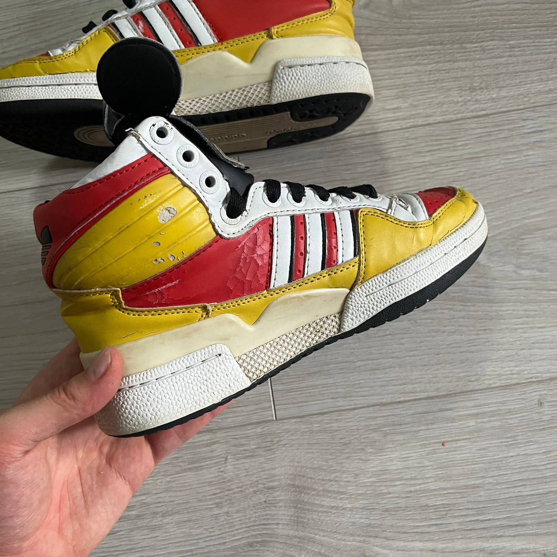 Yellow, red, and white sneaker with black accents held by a hand on a wooden floor.