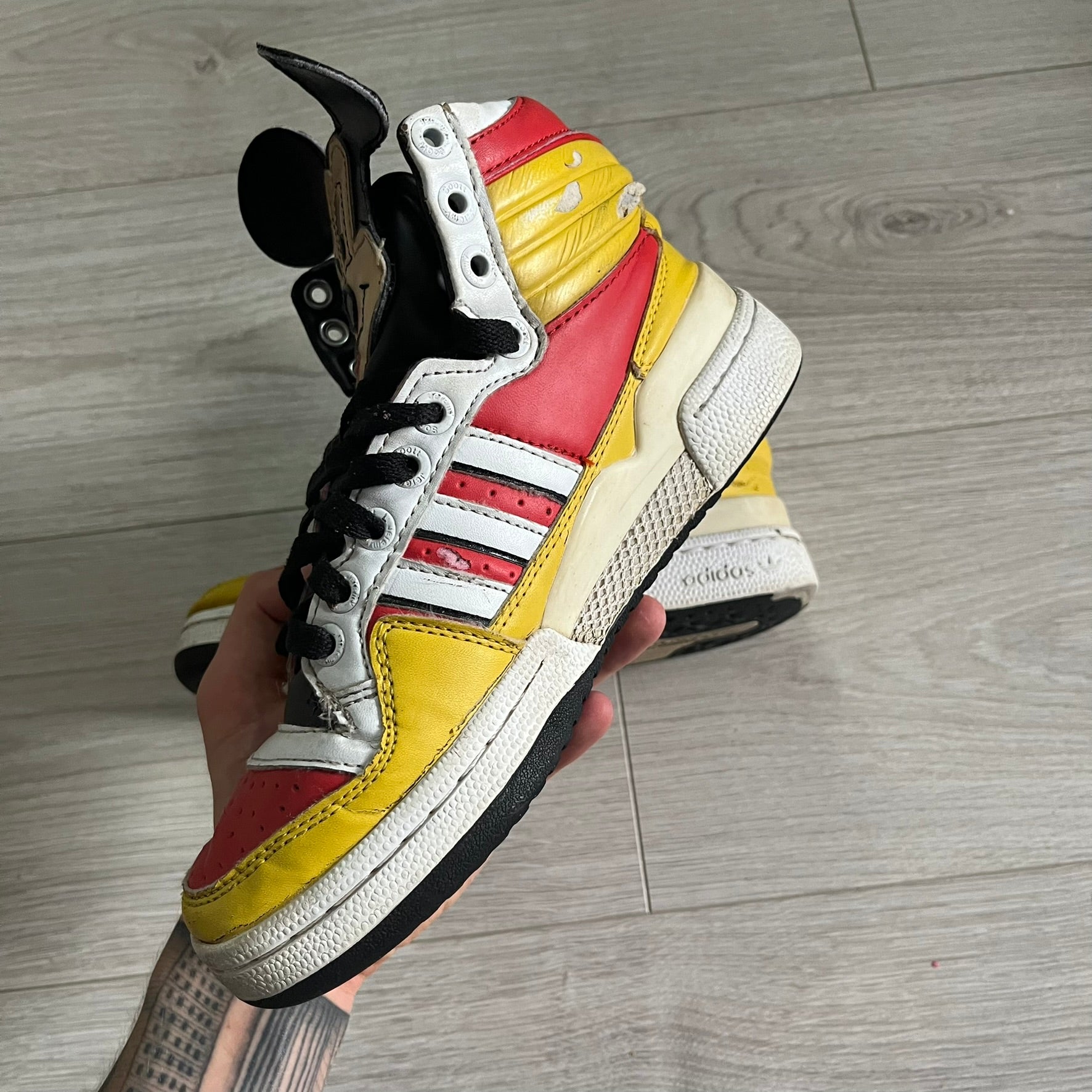 Yellow, red, and white sneaker with black accents held by a hand on a wooden floor.
