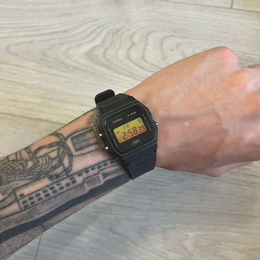 Black Casio digital watch on a wrist with tattoos, on a light wooden surface.