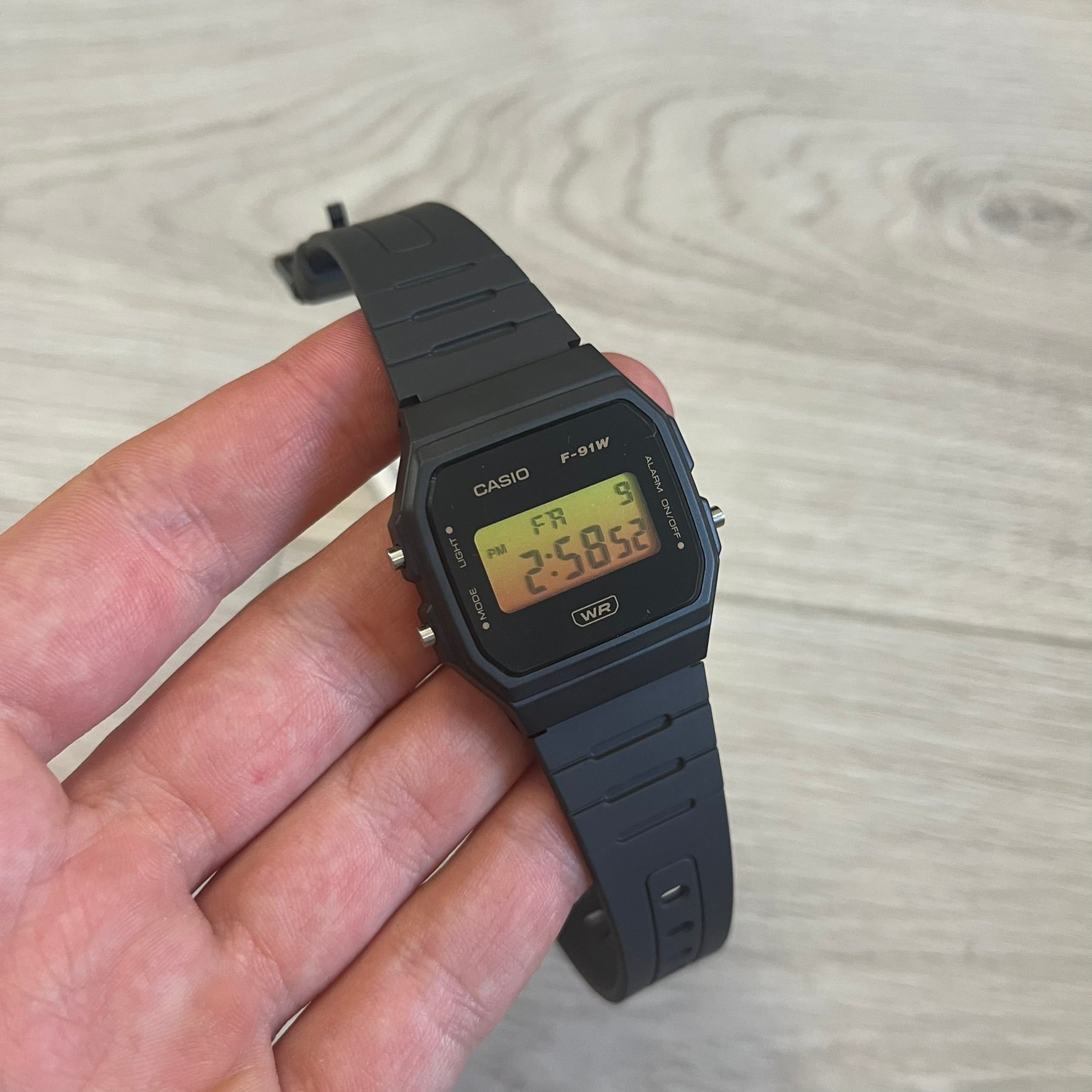 Black Casio digital watch held in a hand on a light wooden surface