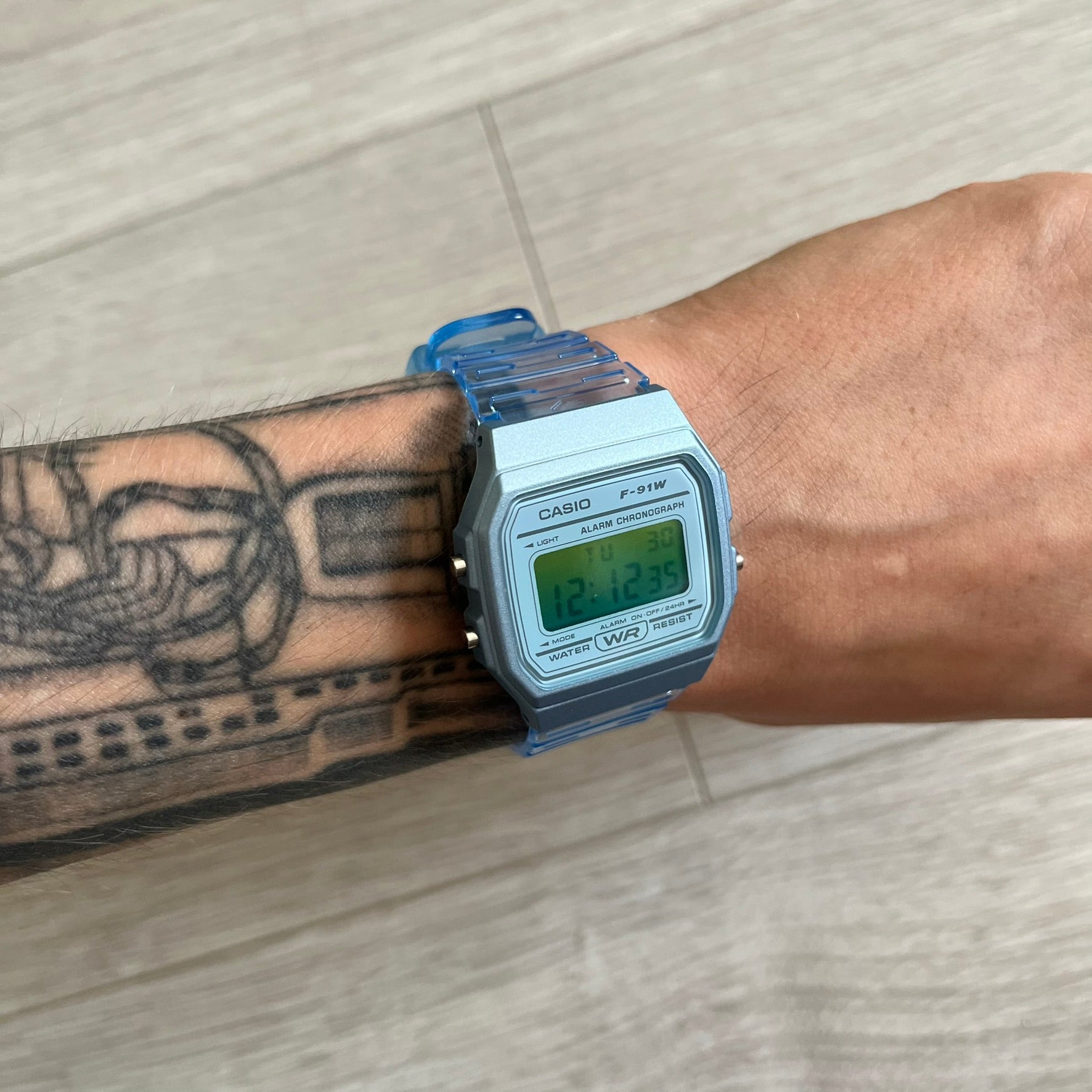 Digital Casio watch on a wrist with tattoos, against a light wooden floor background