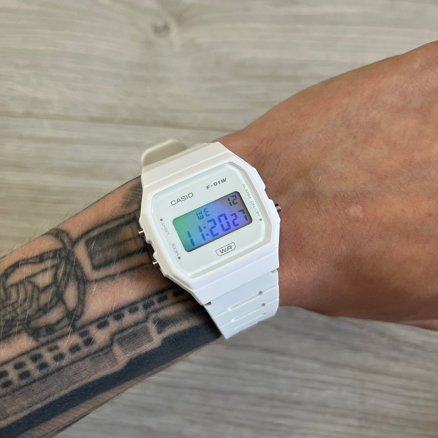 White Casio digital watch on a wrist with tattoos, against a wooden surface background.