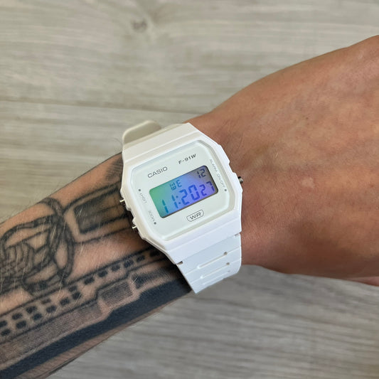 White Casio digital watch on a wrist with tattoos, against a wooden surface background.