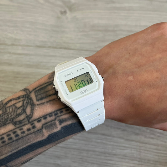 White Casio digital watch on a wrist with tattoos, against a wooden surface background.