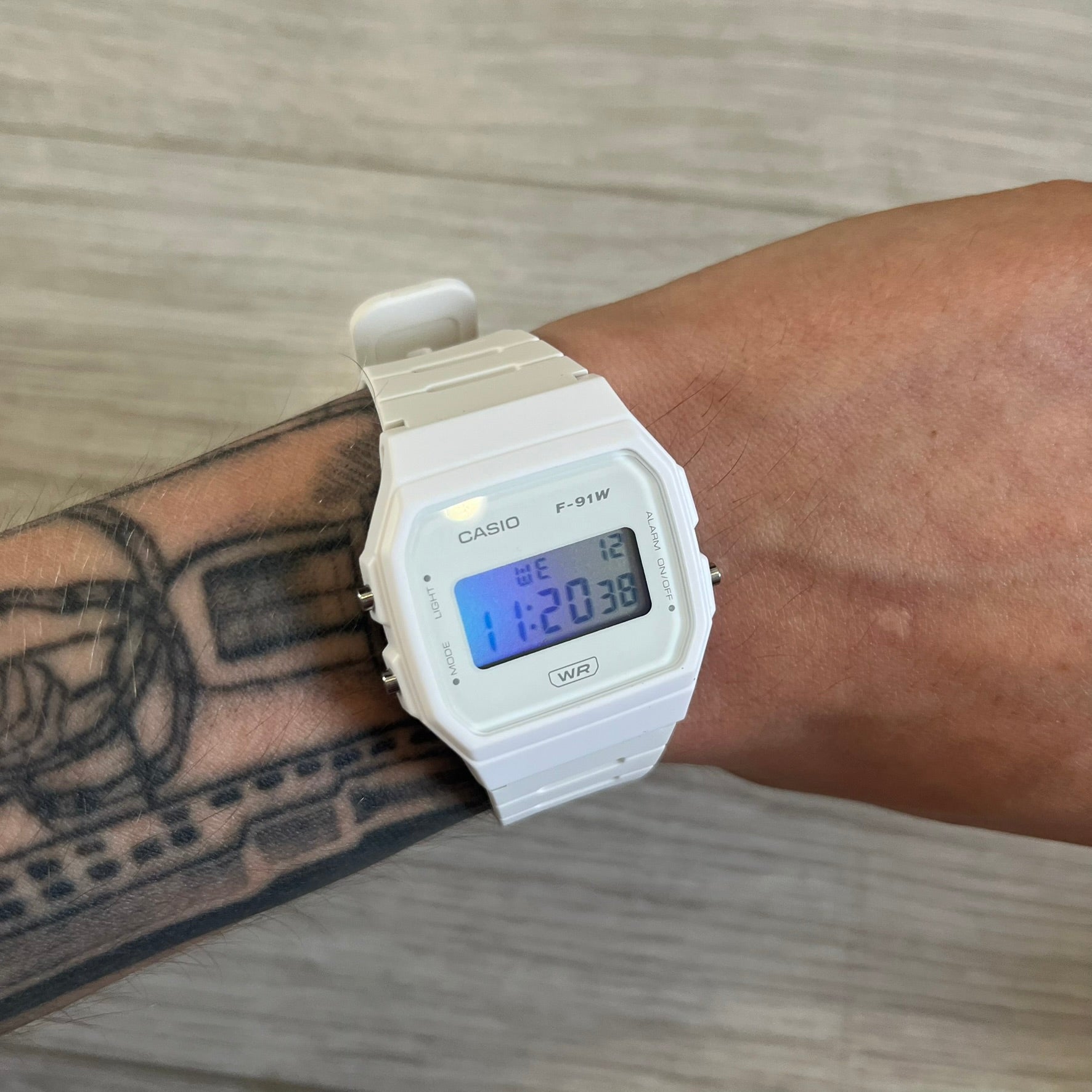 Casio digital watch on a wrist with a tattooed arm, on a wooden surface.
