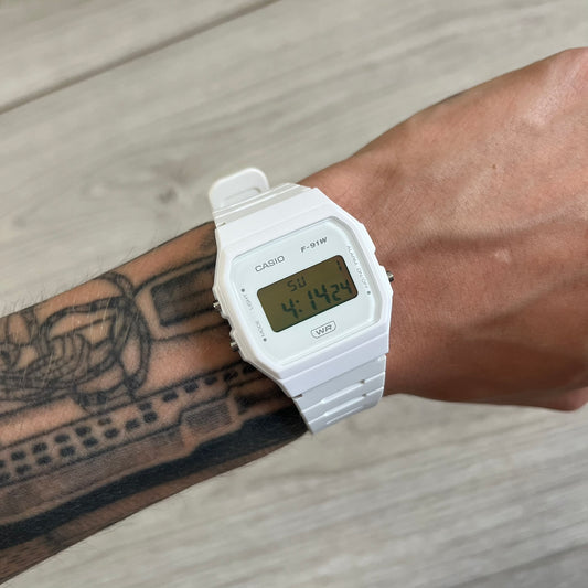 White Casio digital watch on a wrist with tattoos, on a light wooden surface.
