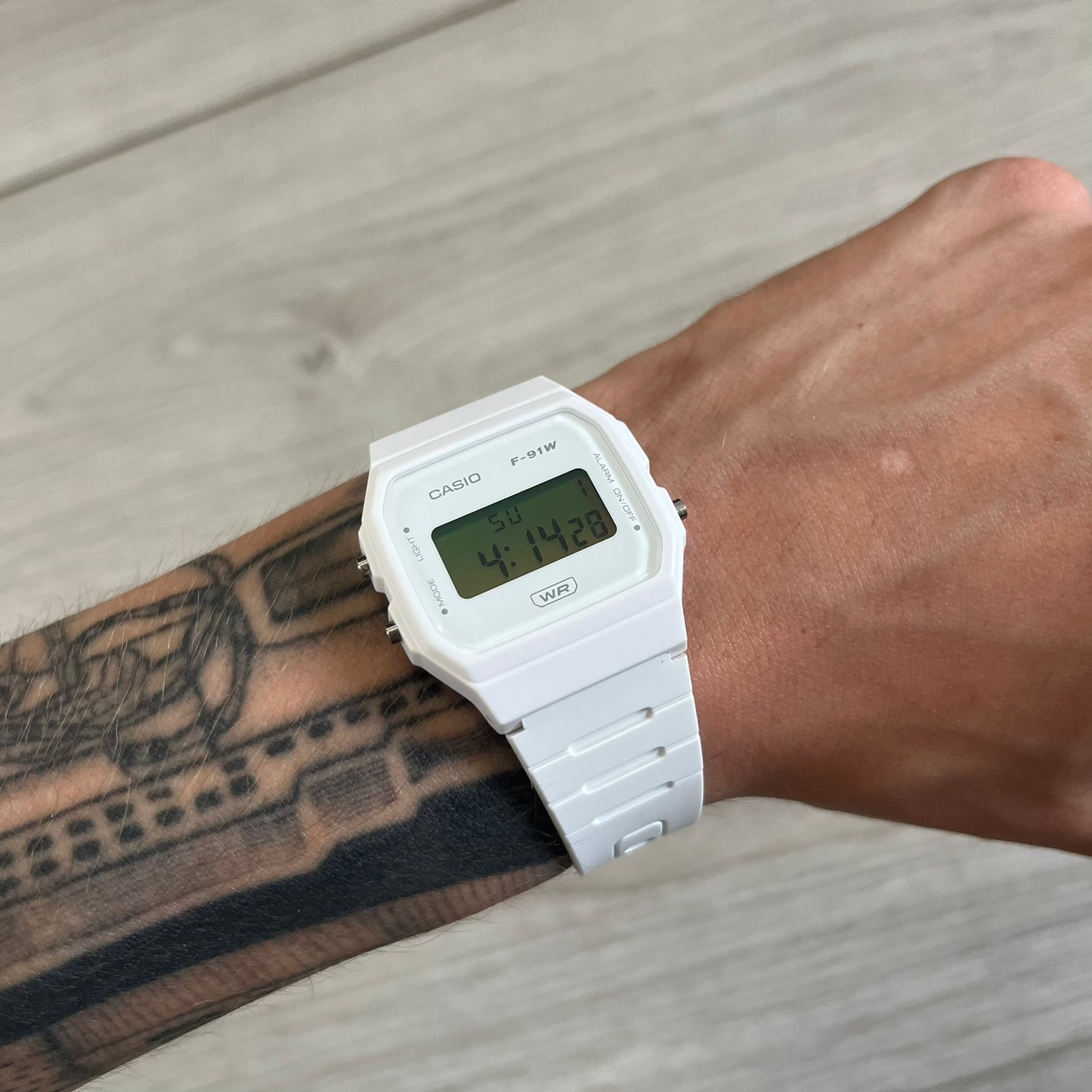 White Casio digital watch on a wrist with tattoos, against a wooden surface.