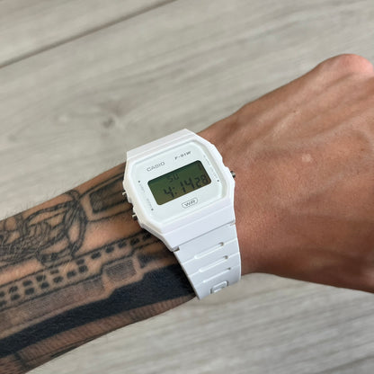 White Casio digital watch on a wrist with tattoos, against a wooden surface.