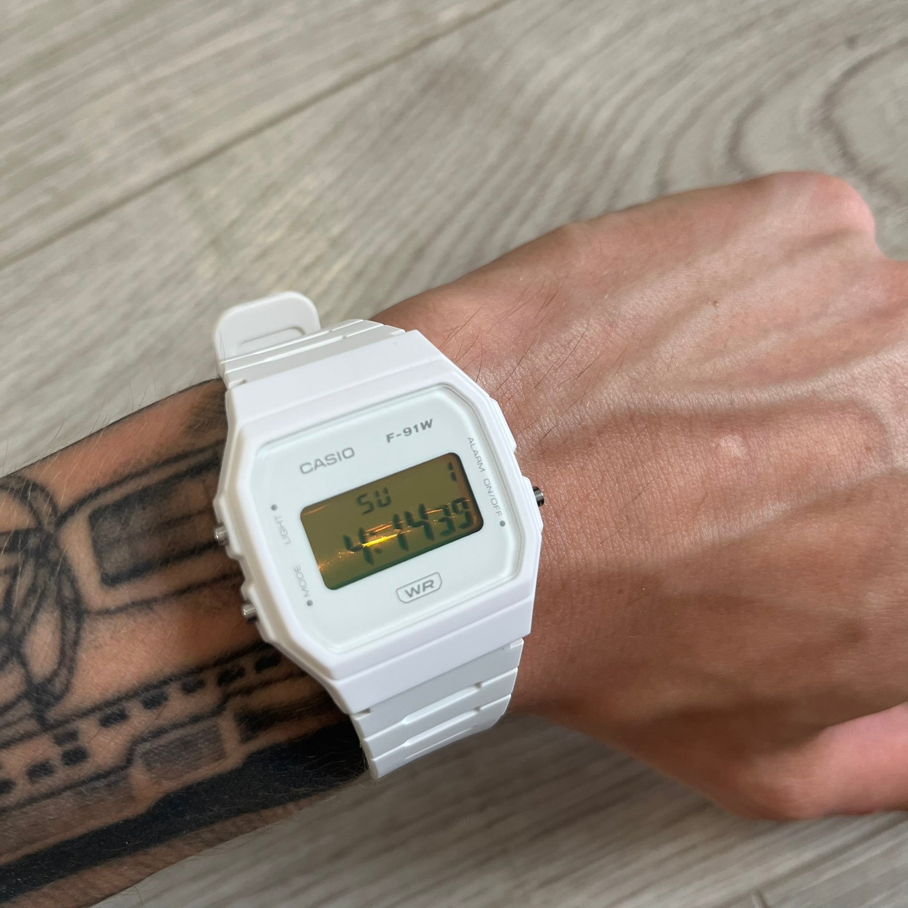 White Casio digital watch on a wrist with a tattooed arm, on a wooden surface.