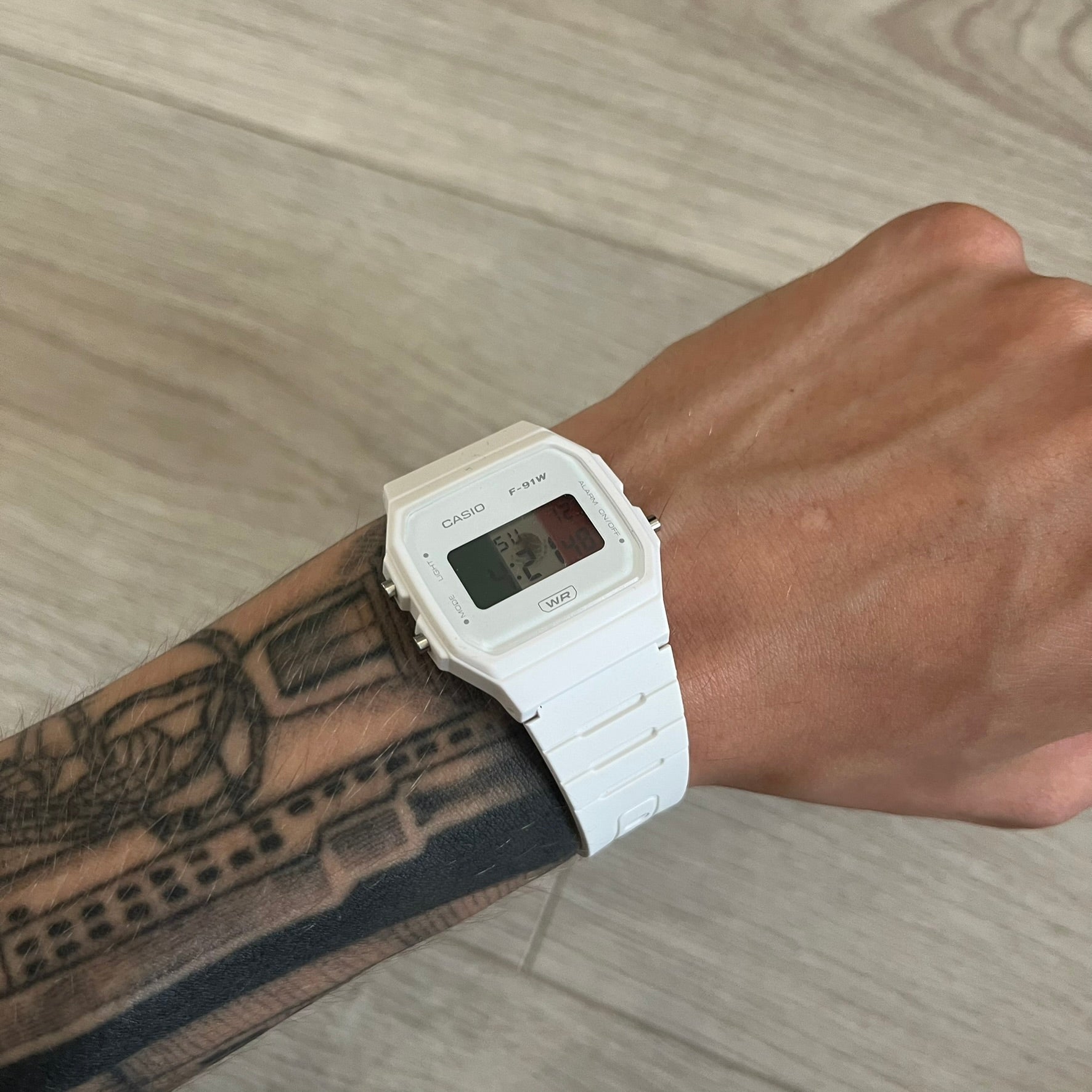 White Casio digital watch on a wrist with tattoos, against a wooden floor background