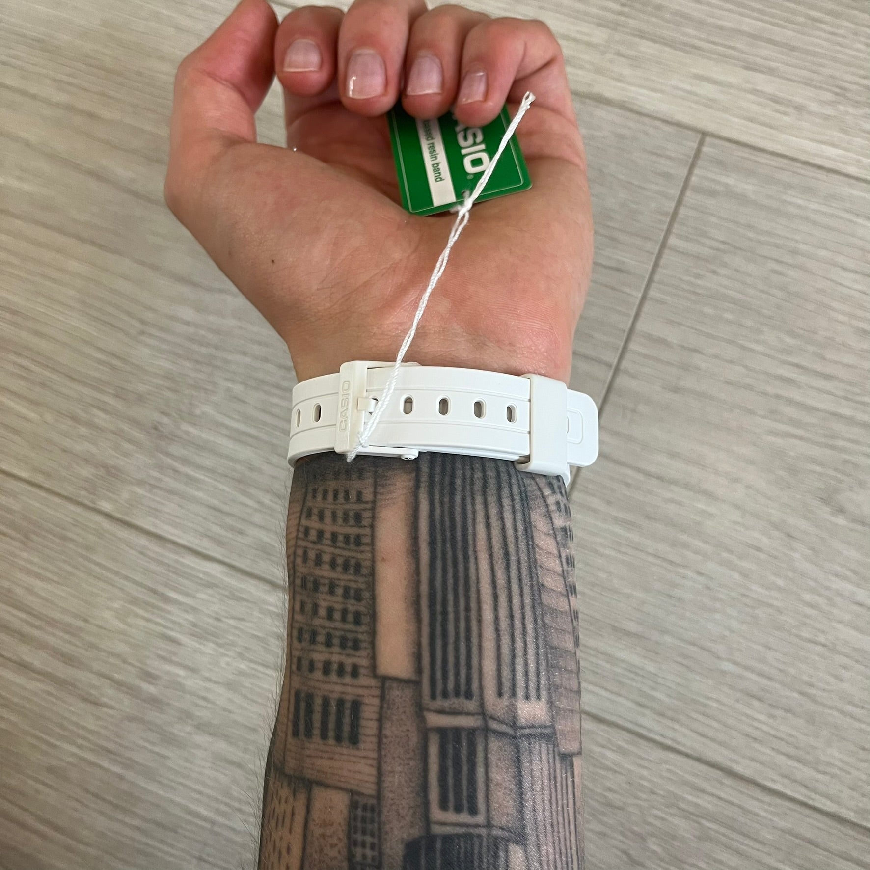 Hand wearing a white watch with a tattooed arm on a wooden floor background