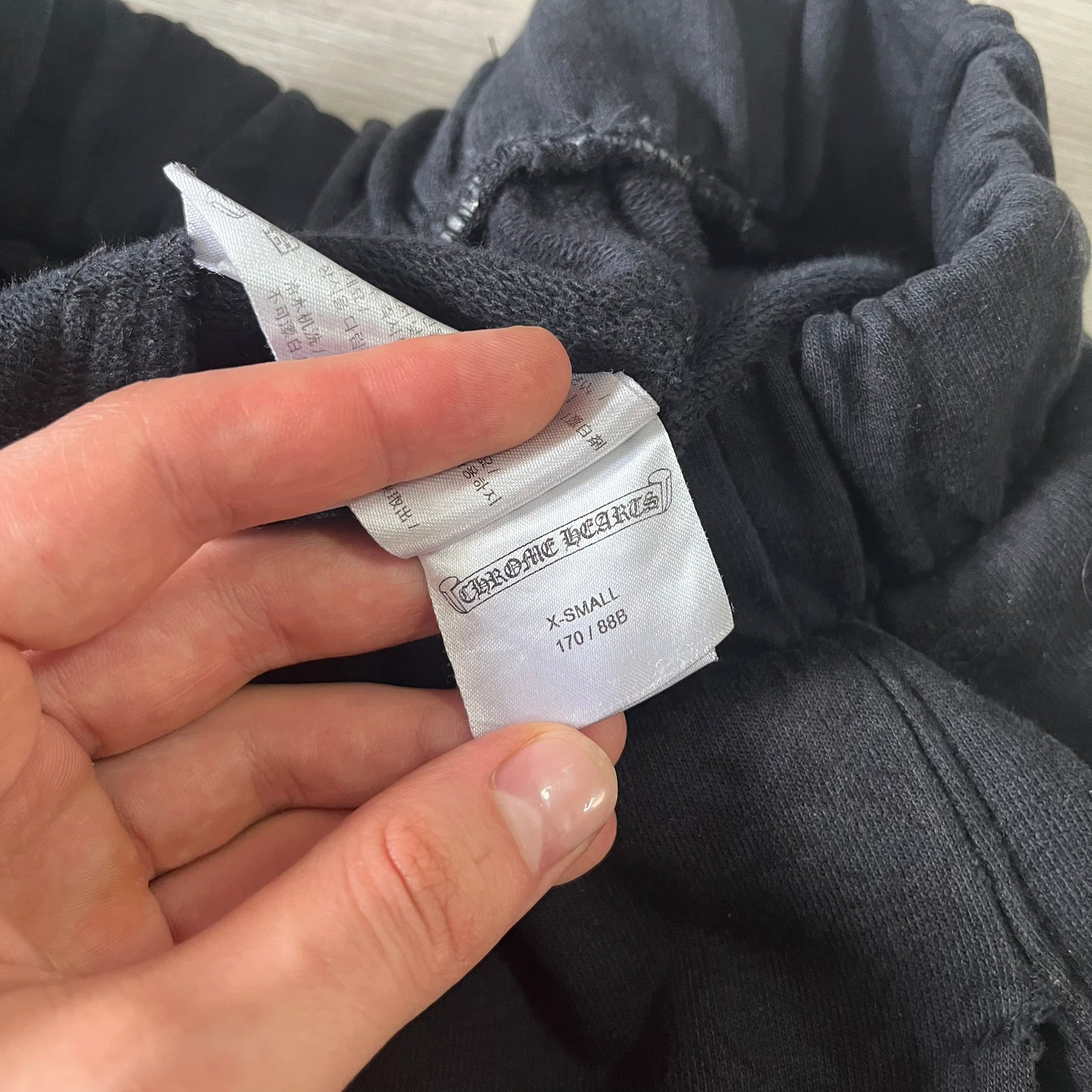 Hand holding a label on a piece of dark gray fabric, with 'Chrome Hearts' branding.