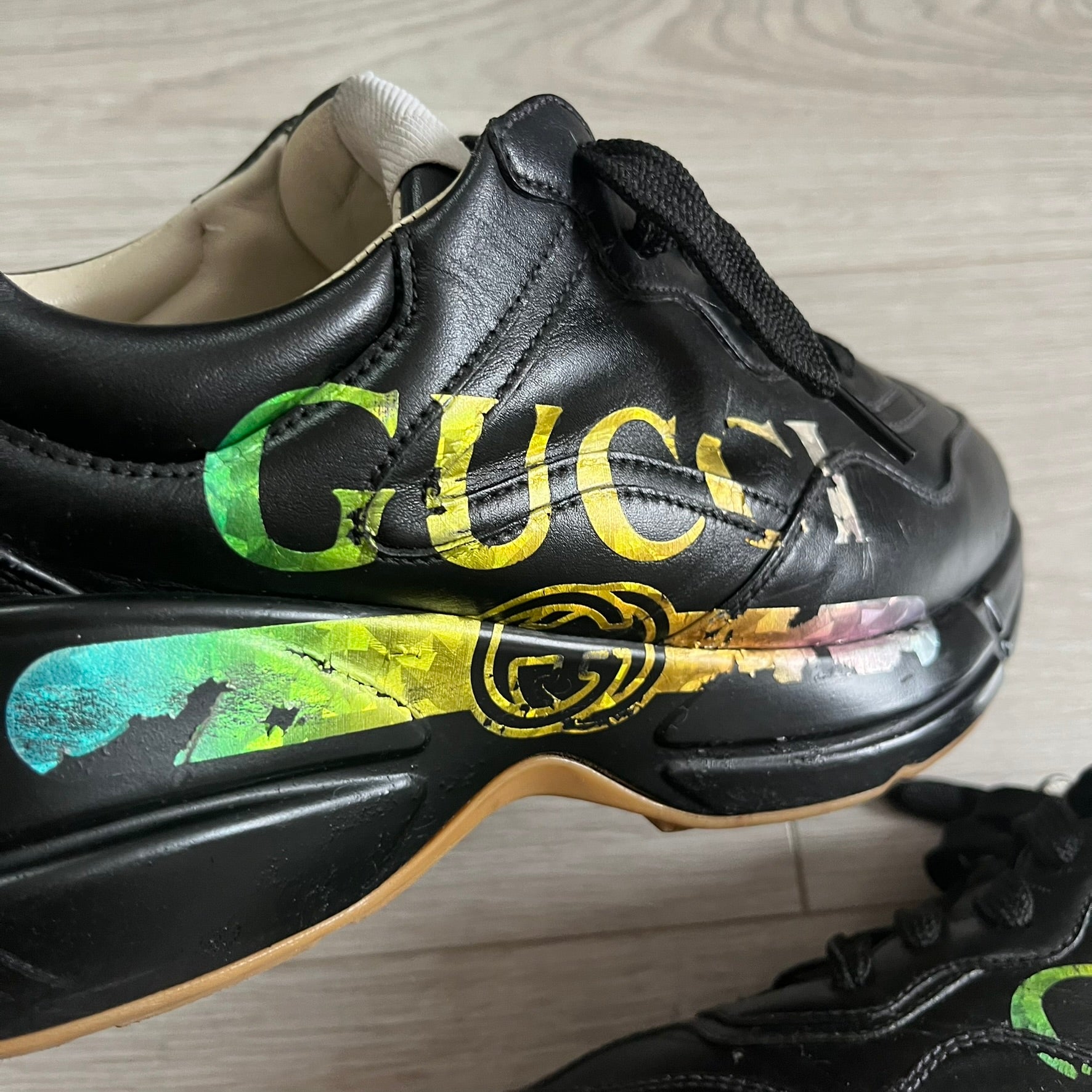 Black sneaker with colorful 'Gucci' logo on a light wooden floor.