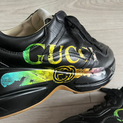 Black sneaker with colorful 'Gucci' logo on a light wooden floor.