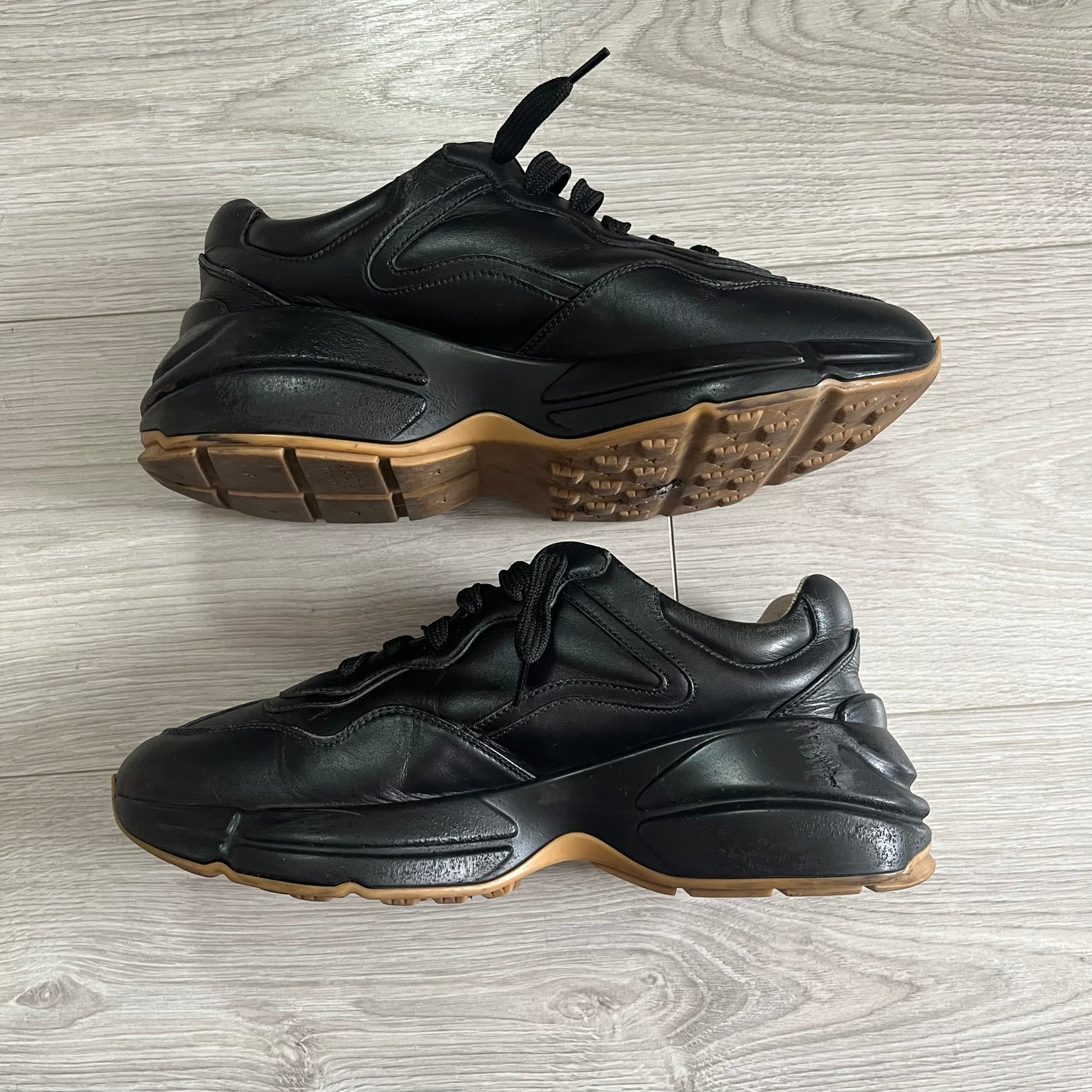 Pair of black sneakers with brown soles on a light wooden floor.