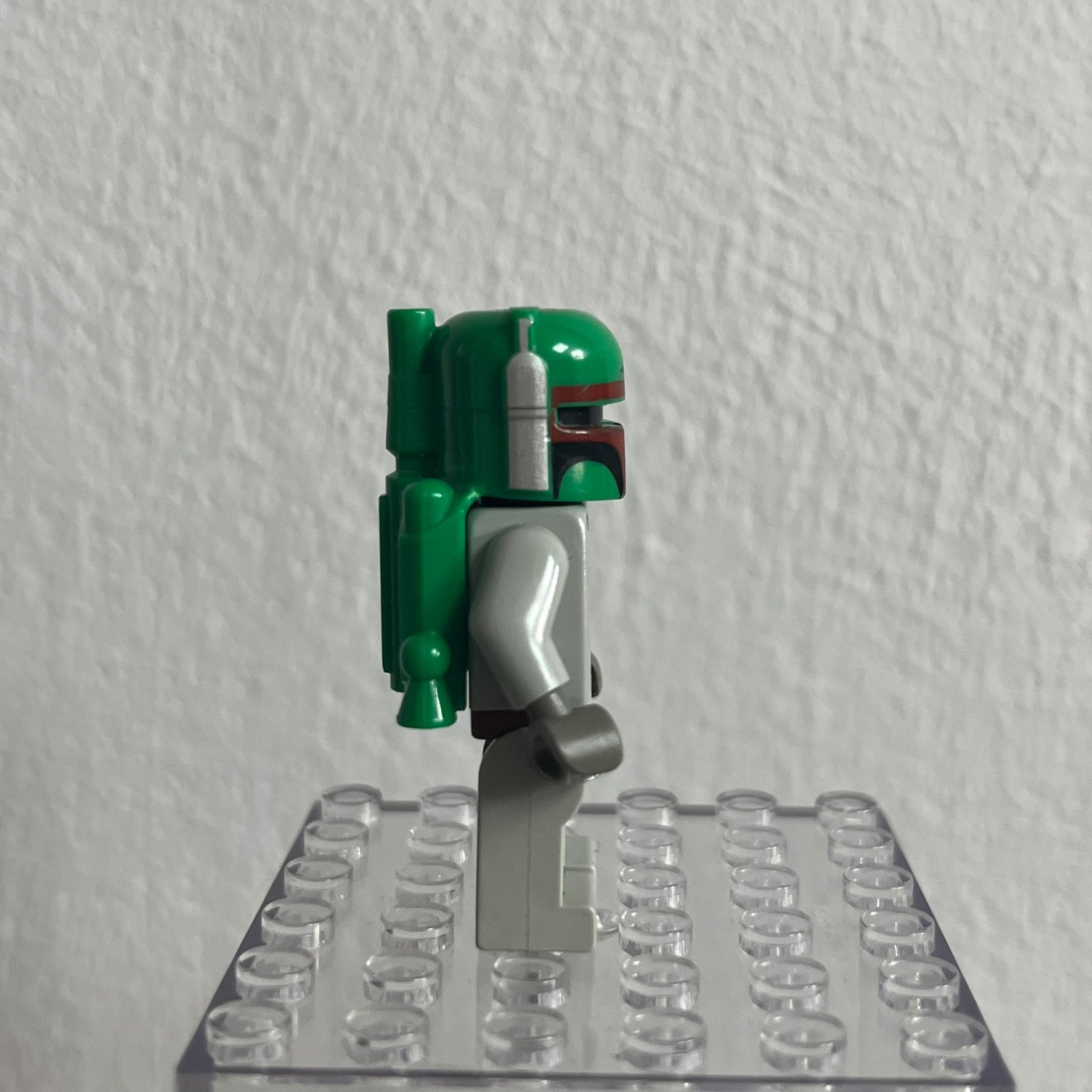 Green and gray Lego figure on a clear baseplate against a white wall