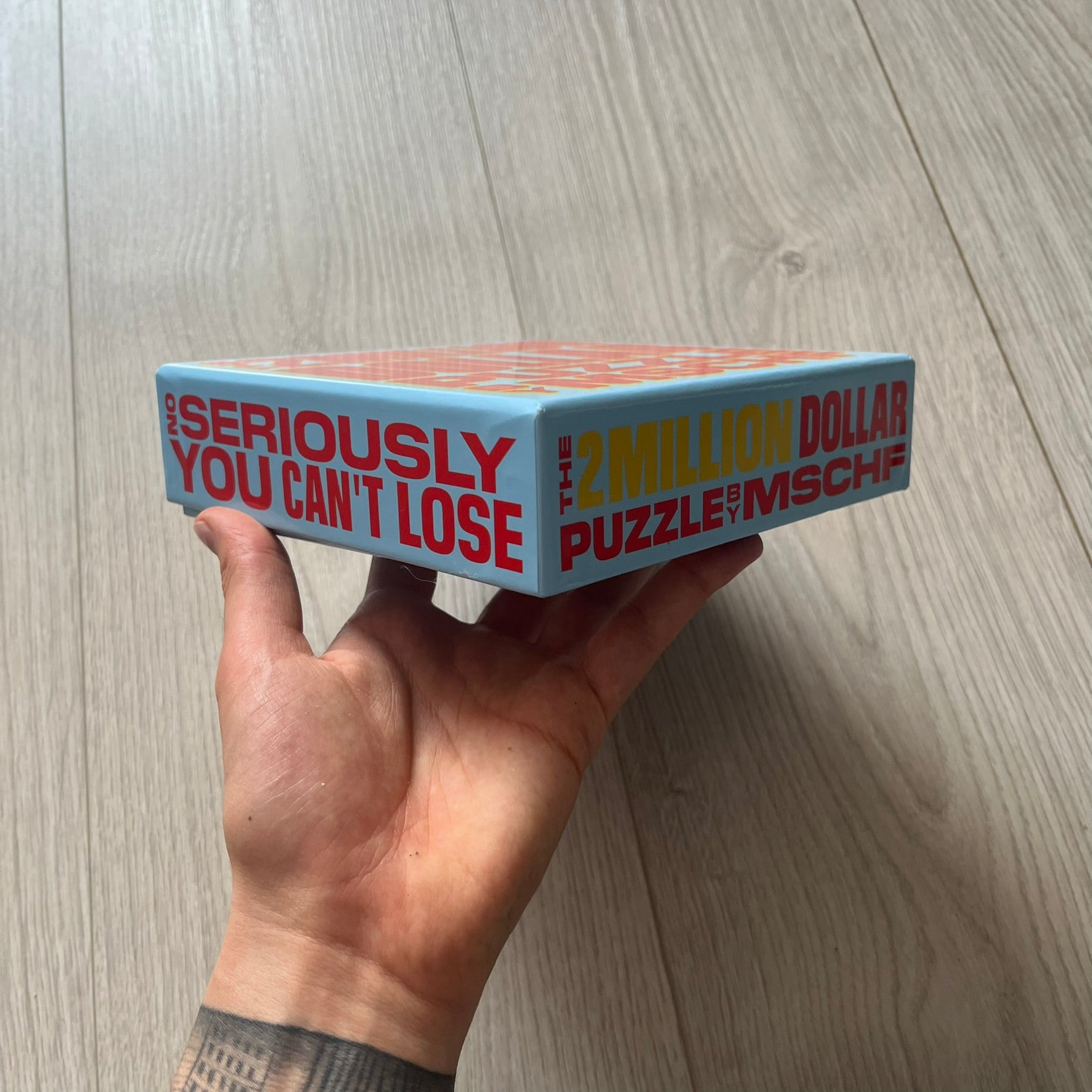 Hand holding a box of '2 Million Dollar Puzzle Mischief' on a wooden floor.