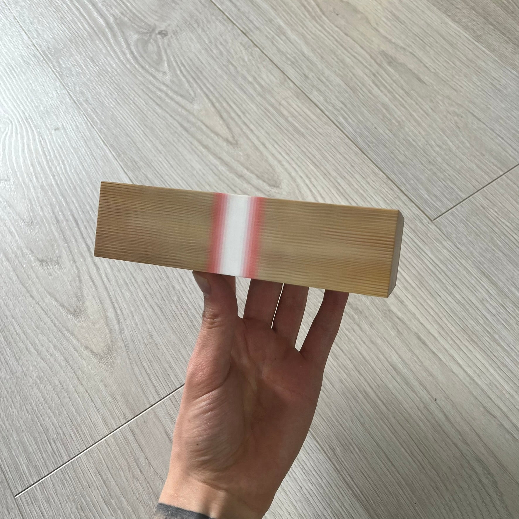 Hand holding a wooden block with a red stripe on a light wood floor.
