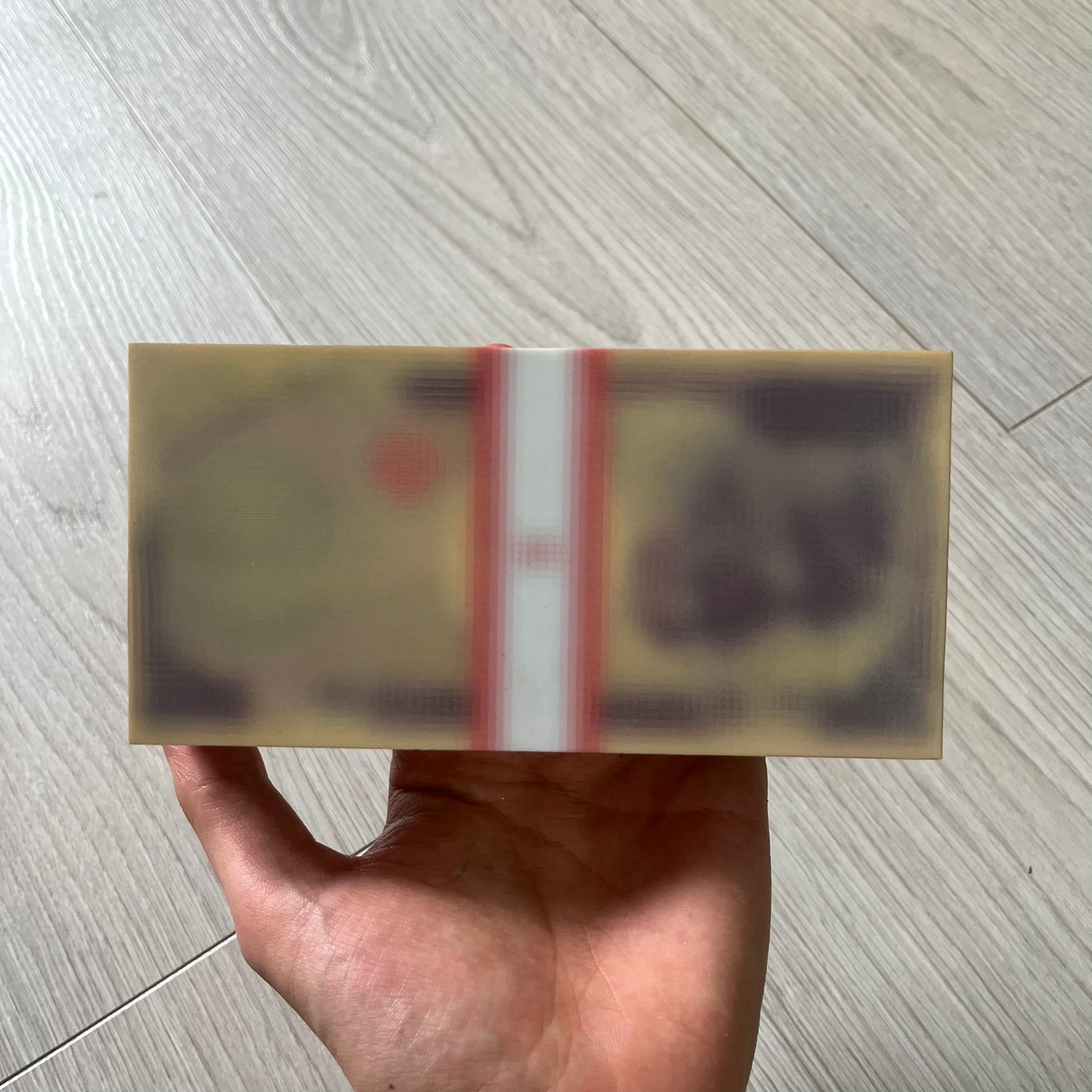 Hand holding a transparent box with a red label on a light wooden floor.