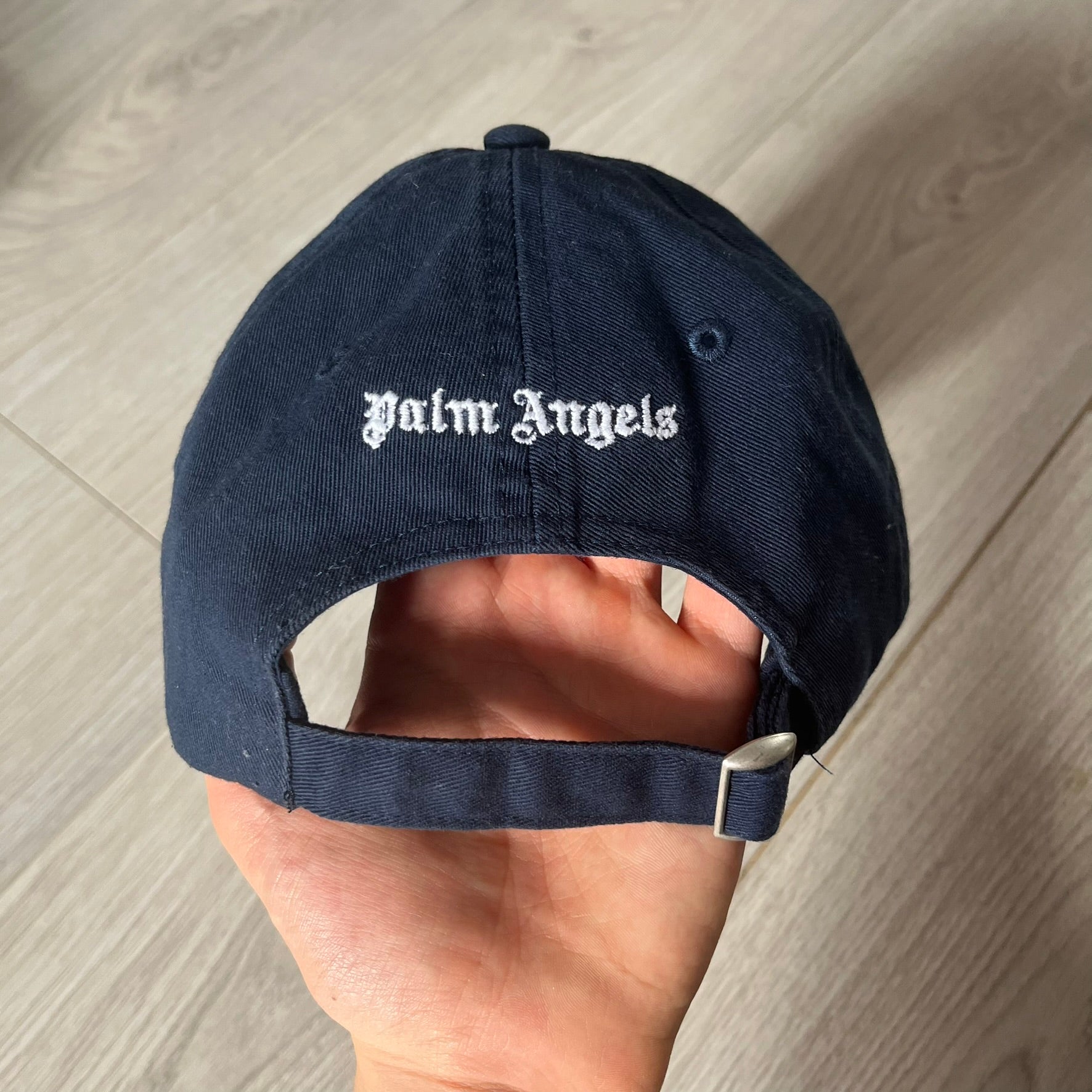 Navy blue cap with 'Palm Angels' logo held by a hand on a wooden surface