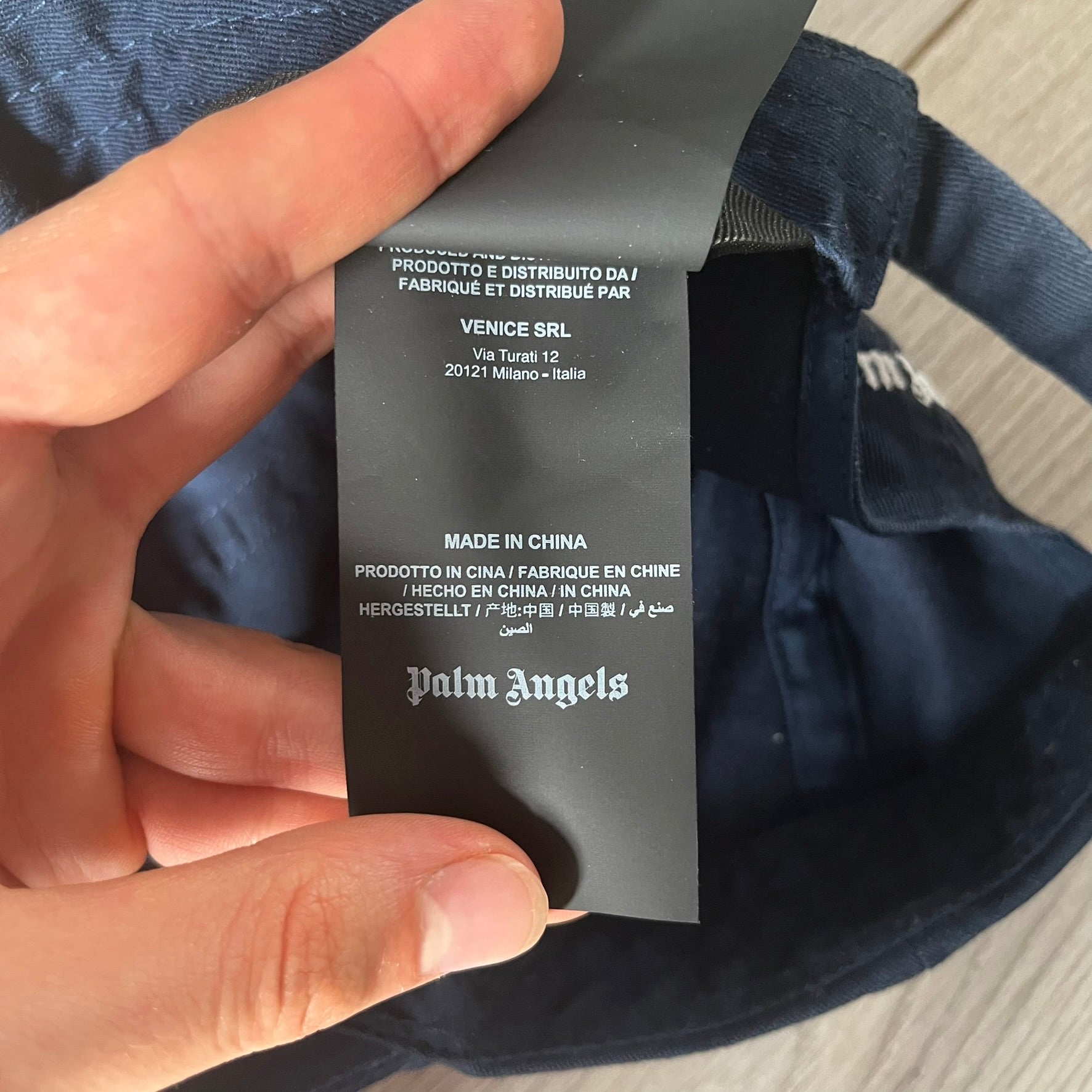 Black label with text and 'Palm Angels' branding held by a hand against a dark fabric background.