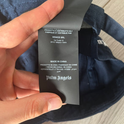 Black label with text and 'Palm Angels' branding held by a hand against a dark fabric background.